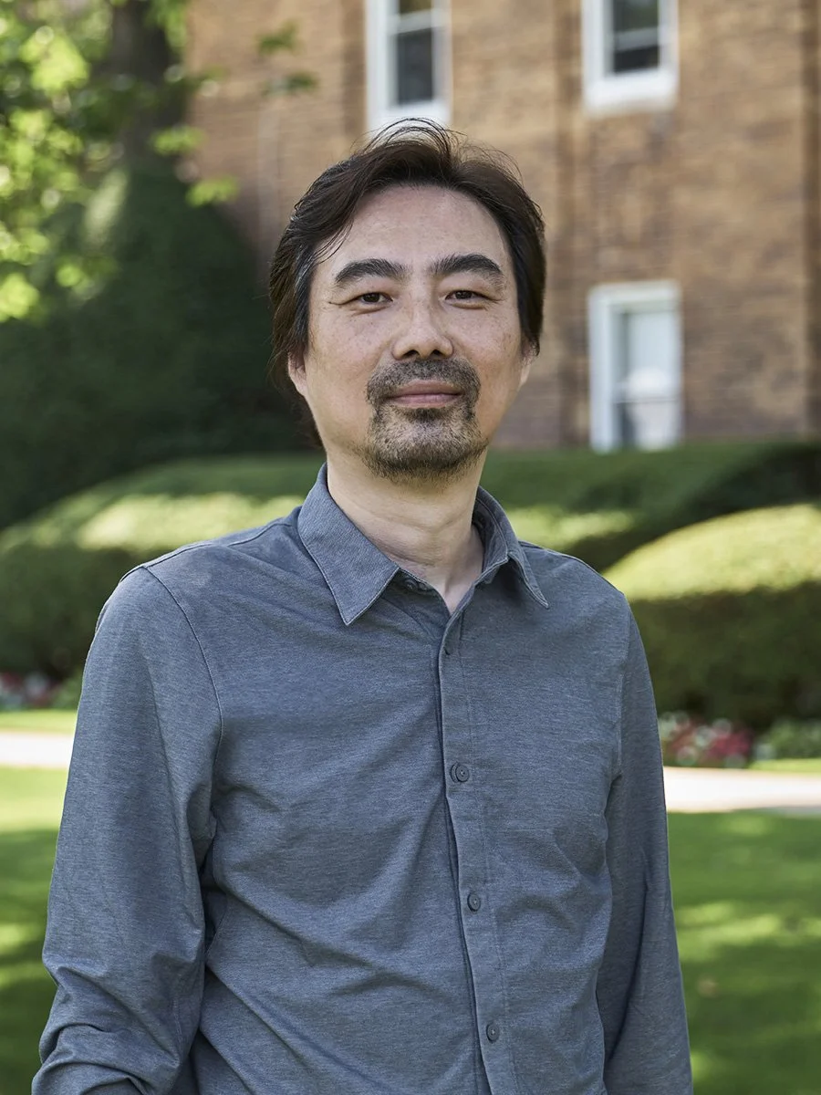 A man with dark hair, a beard, and a mustache stands outdoors in front of greenery and a brick building, wearing a gray button-up shirt.