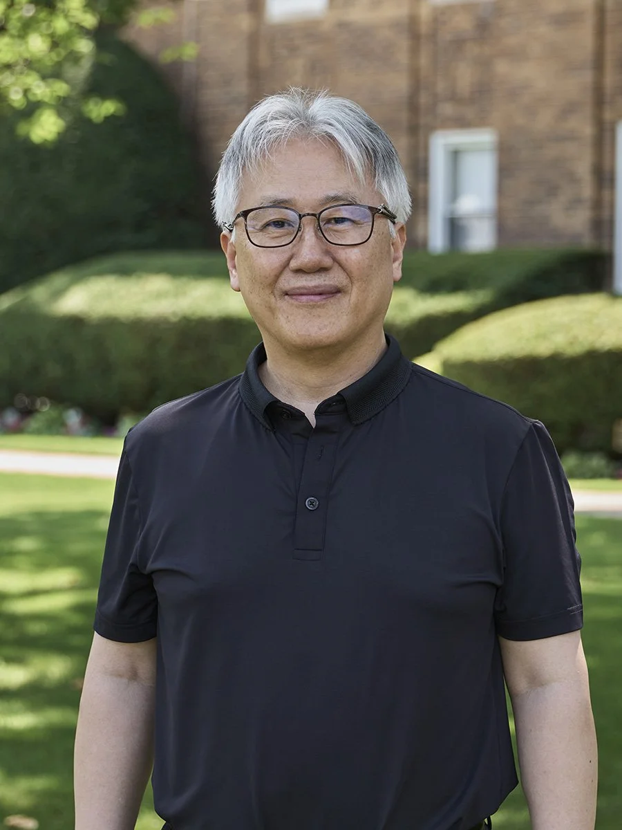A man with gray hair and glasses wearing a black polo shirt standing outdoors in front of a house with a well-kept yard and trees.