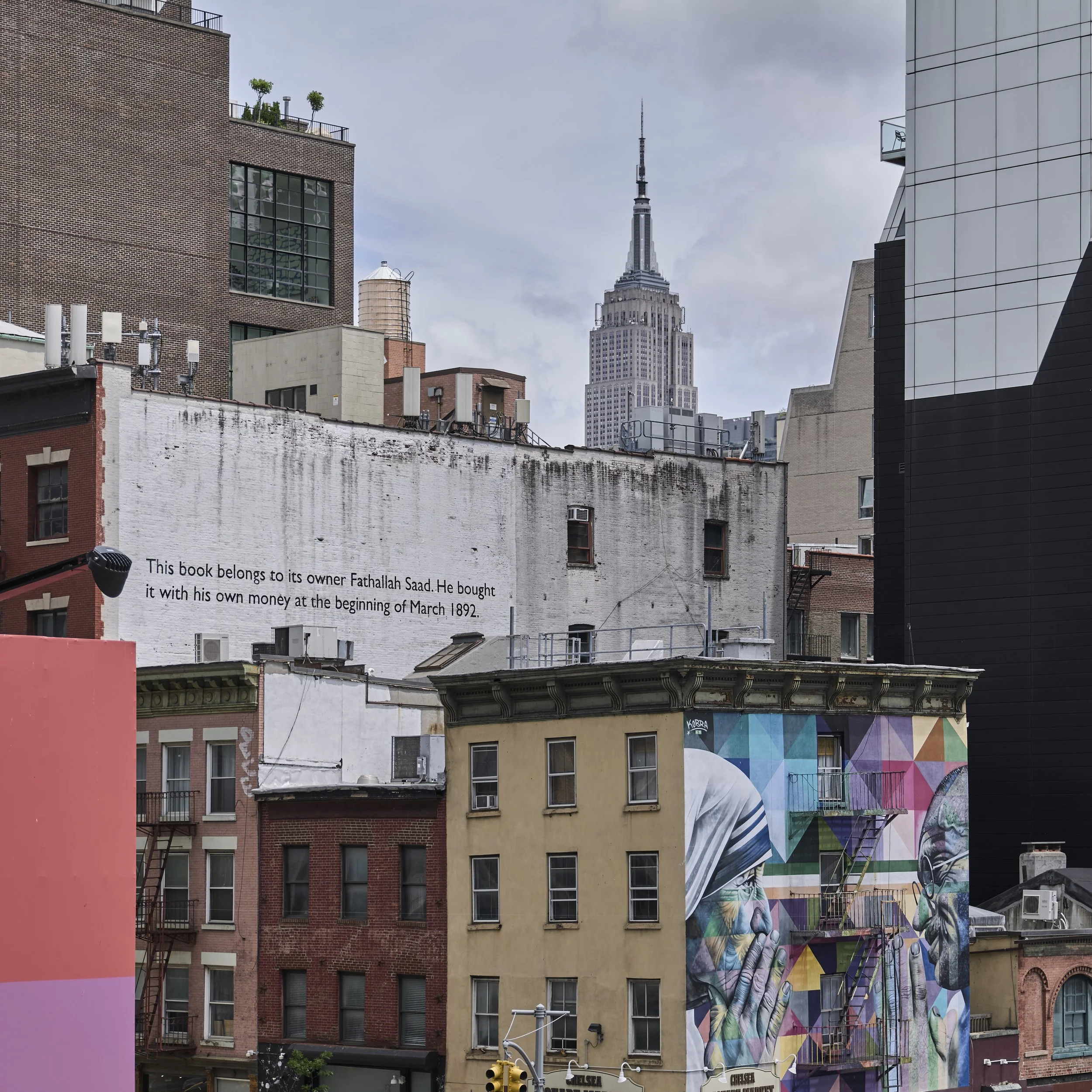Cityscape of New York City with the Empire State Building in the background. Foreground features various buildings, including one with a mural of two hands holding a pair of glasses, and a white building with a quote about a book belonging to its owner.