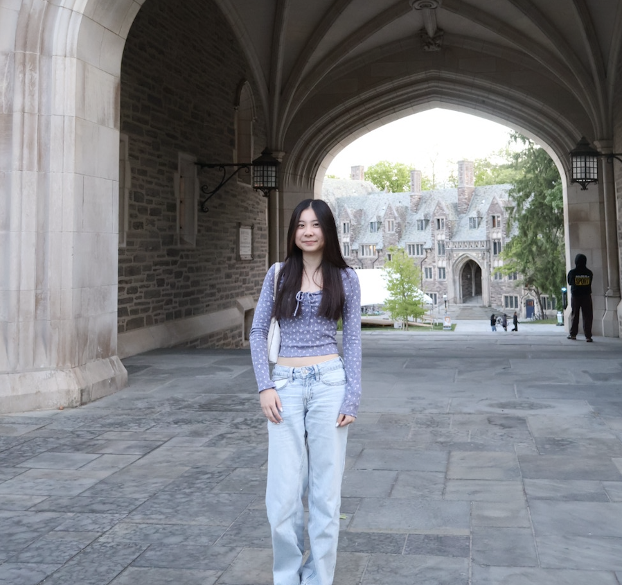 A young woman with long dark hair standing under a stone archway on a university campus. She is wearing a purple polka-dotted sweater and light blue jeans. In the background, there is a historic stone building with a large arched entrance and a few people walking around.