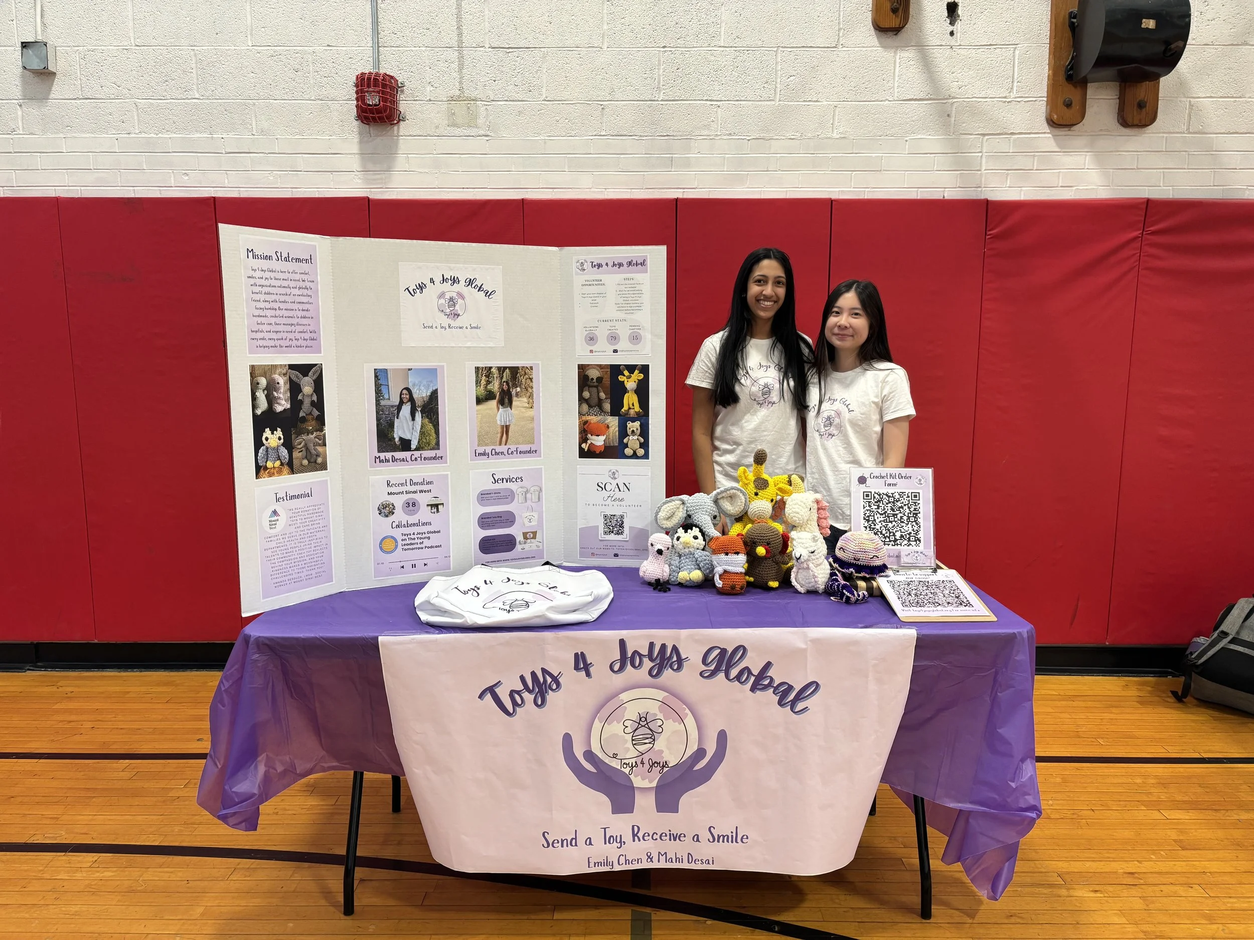 Two young women standing behind a table with a display board and handmade crochet toys, at a community event or fair. The display board features information and photos about Toys 4 Joys Global, an organization that sends toys to children. Decorated in purple and white, the table also has a large banner with the organization's logo and slogan, and various crocheted toys are arranged on the table.