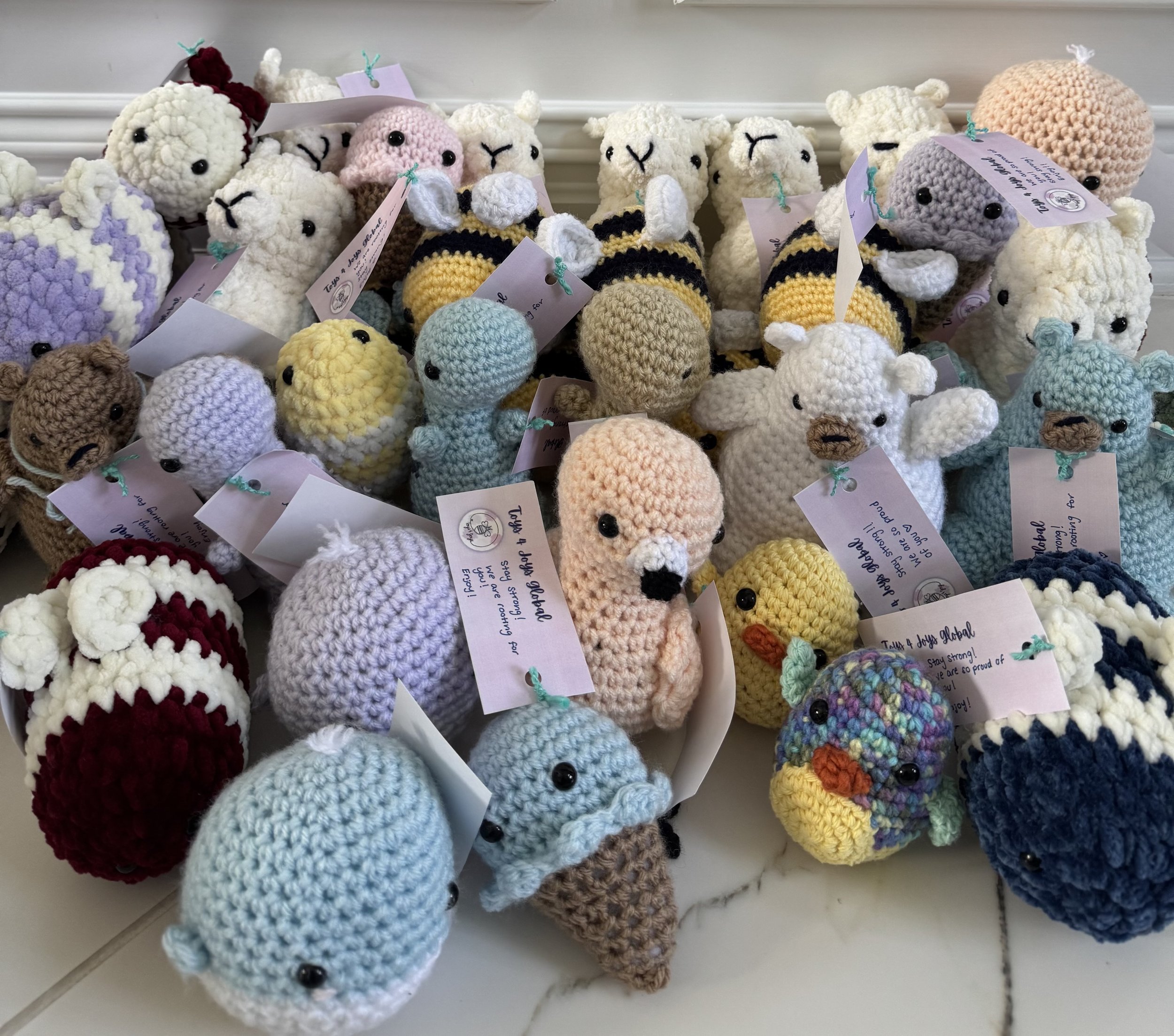 Collection of handmade crochet plush toys including sheep, bees, ducks, and other animals, each with attached tags.