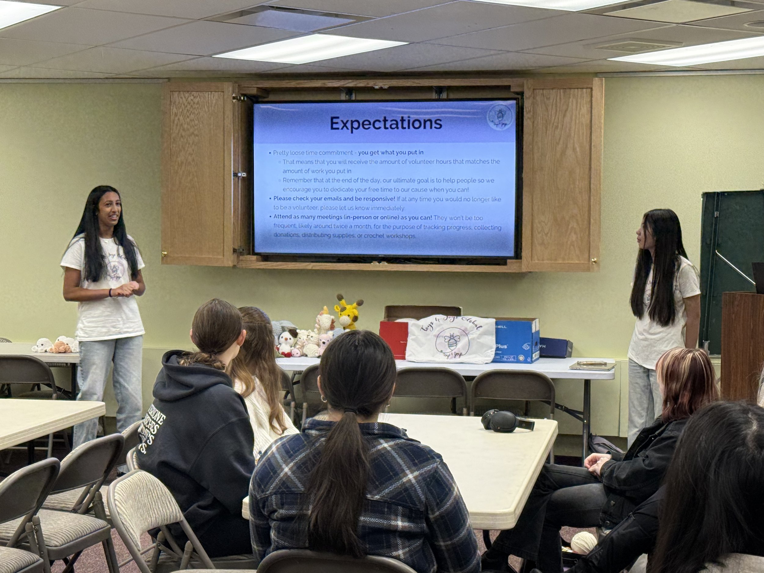 Two women presenting to an audience in a room with a large screen displaying a slide titled "Expectations" with bullet points about volunteer work and meeting attendance. There are chairs, tables, and stuffed toys on the tables in the room.