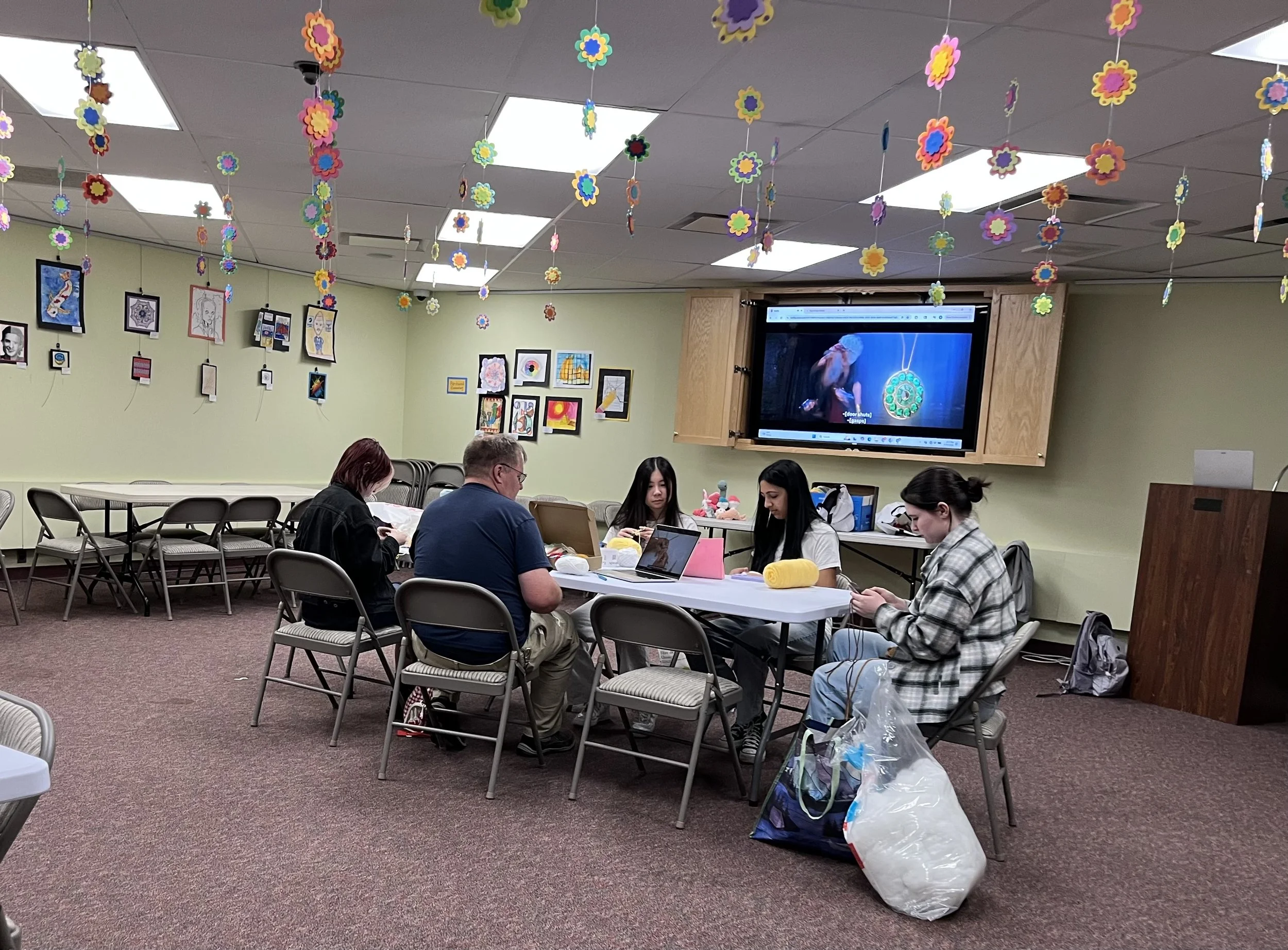 People sitting at tables in a room decorated with hanging paper flower decorations and artwork on the walls, watching a presentation on a large TV screen.