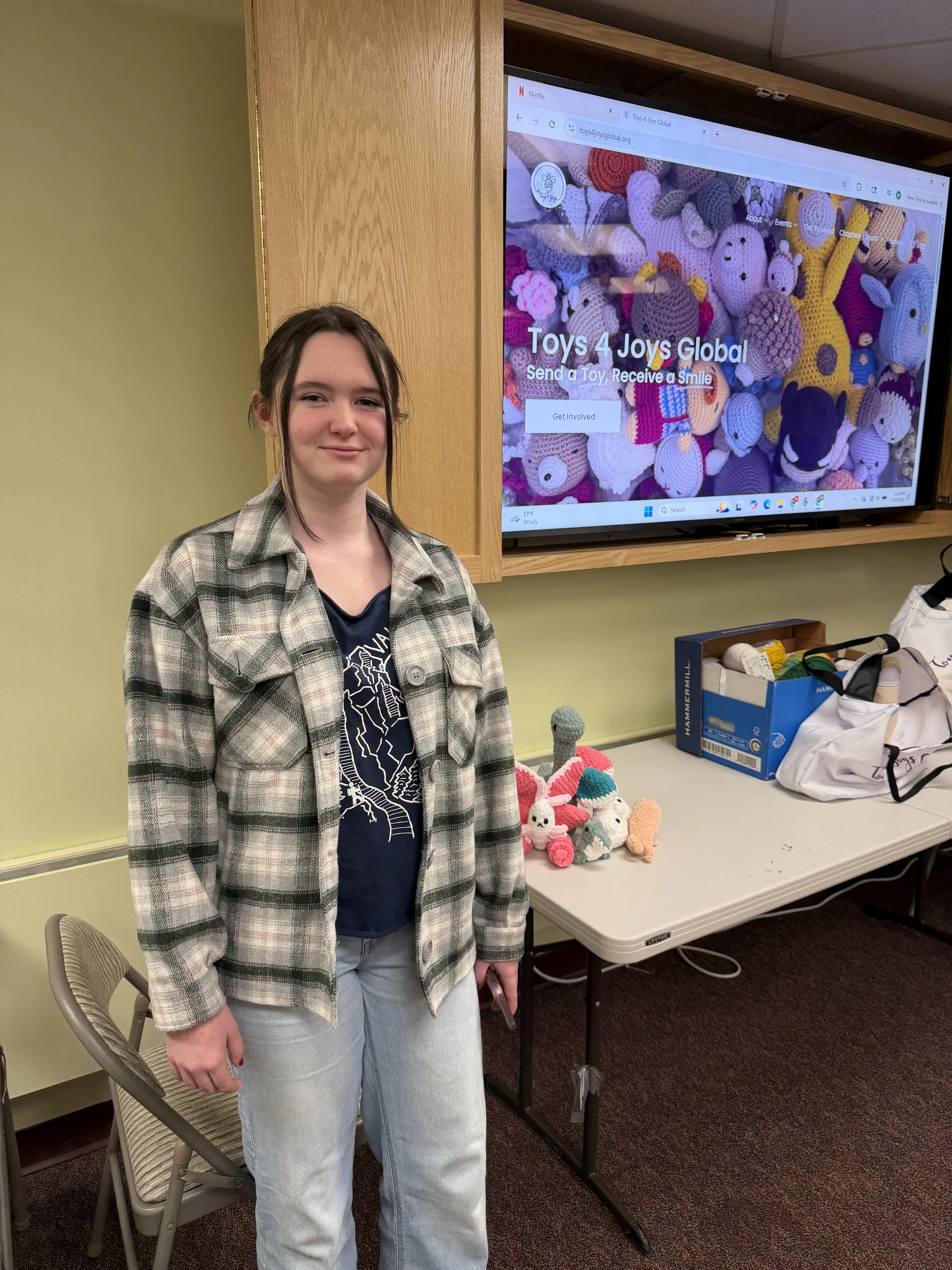 A young girl with long dark hair, wearing a plaid jacket over a dark shirt and light jeans, standing in front of a table with handmade crocheted stuffed animals in bright colors. Behind her is a large screen displaying the homepage of a website called Toys 4 Joys Global, with the slogan 'Send a Toy, Receive a Smile.'