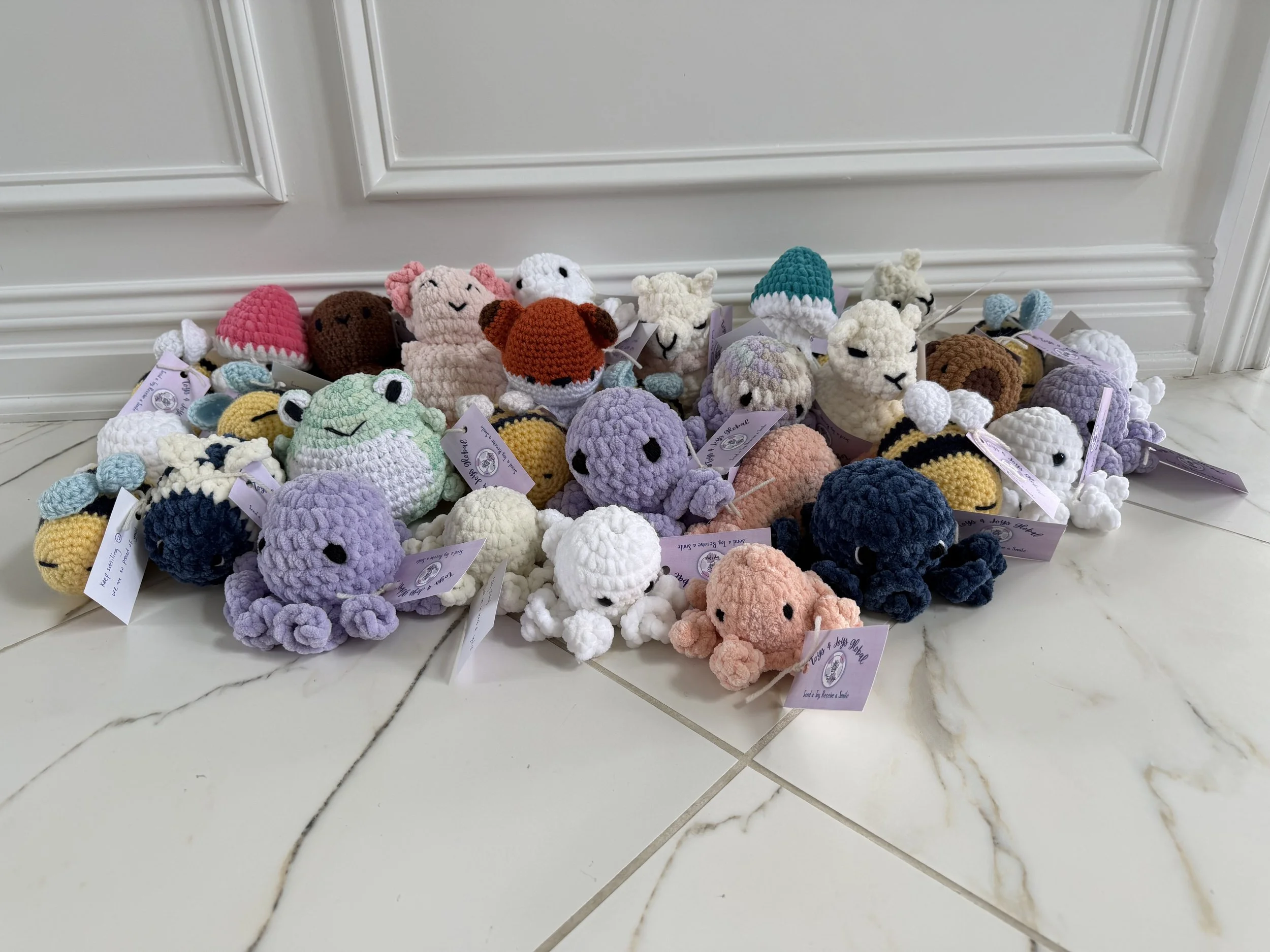 A collection of colorful crocheted stuffed animals, including various animals and characters, arranged on a tiled floor against a white paneled wall.