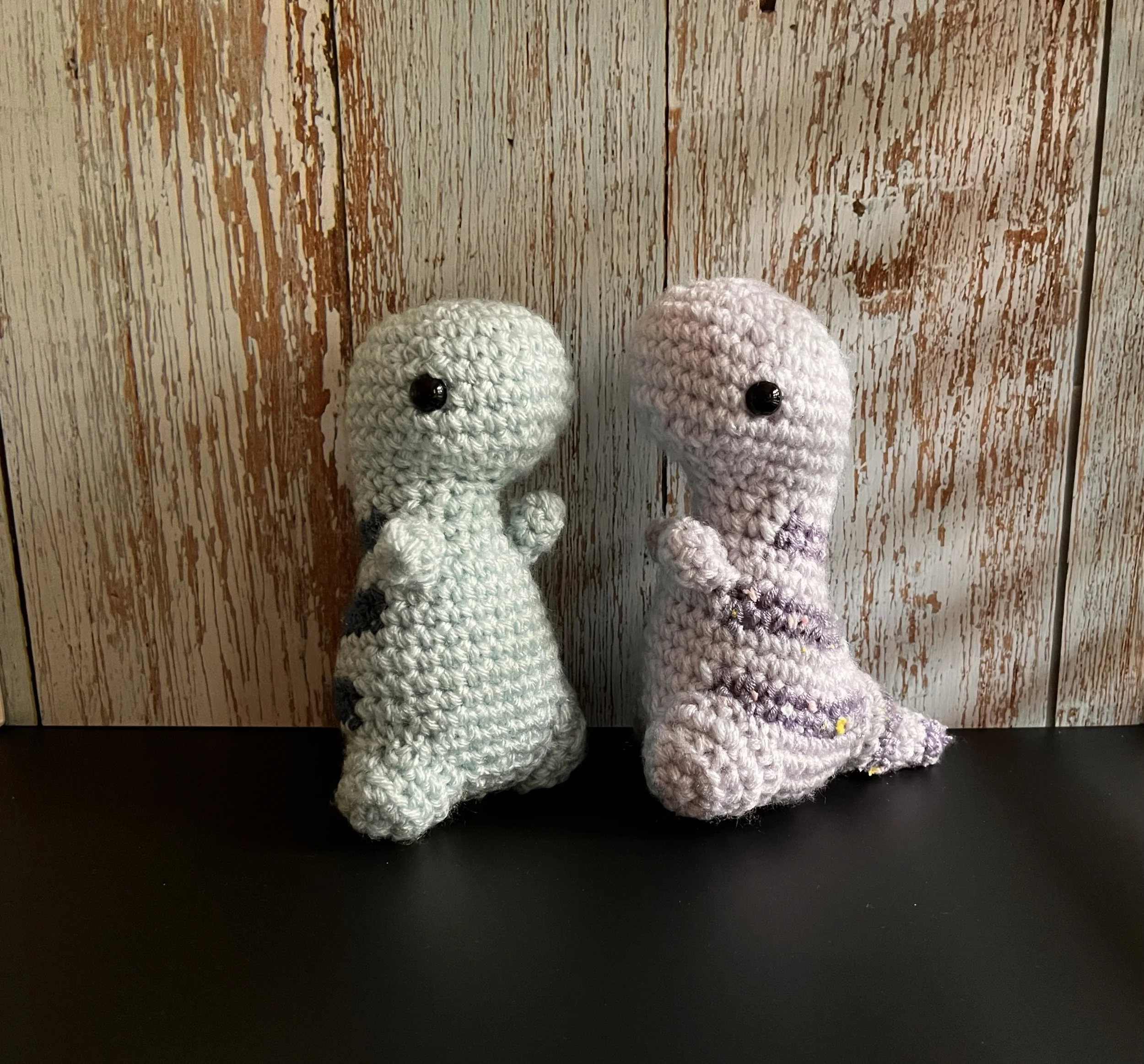 Two crocheted stuffed alien dolls with button eyes, standing on a black surface in front of a wooden textured background.
