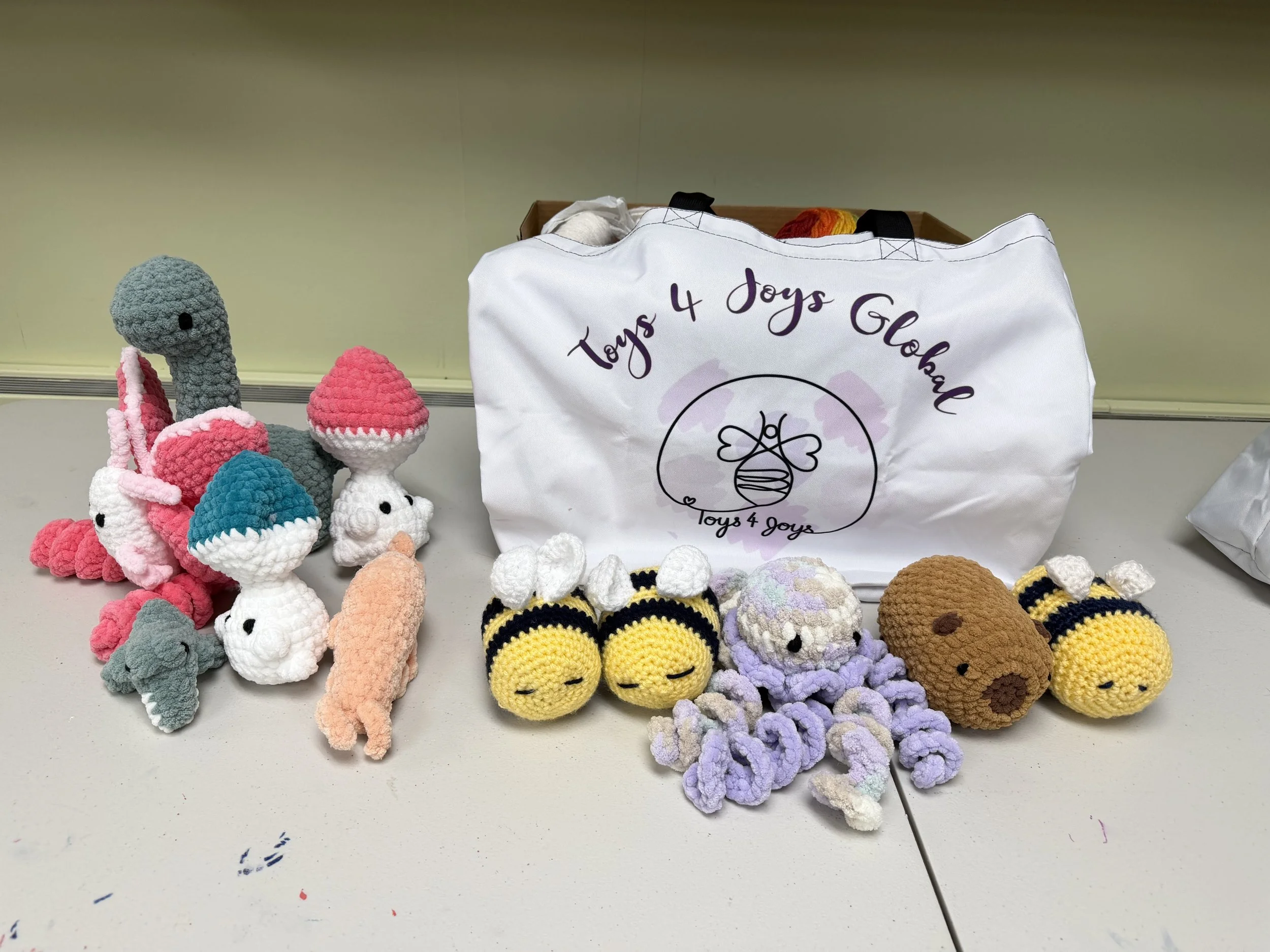 A collection of crocheted toys including bunnies, cats, bees, and other animals arranged on a table, with a white bag labeled 'Toys 4 Joys Global' in the background.