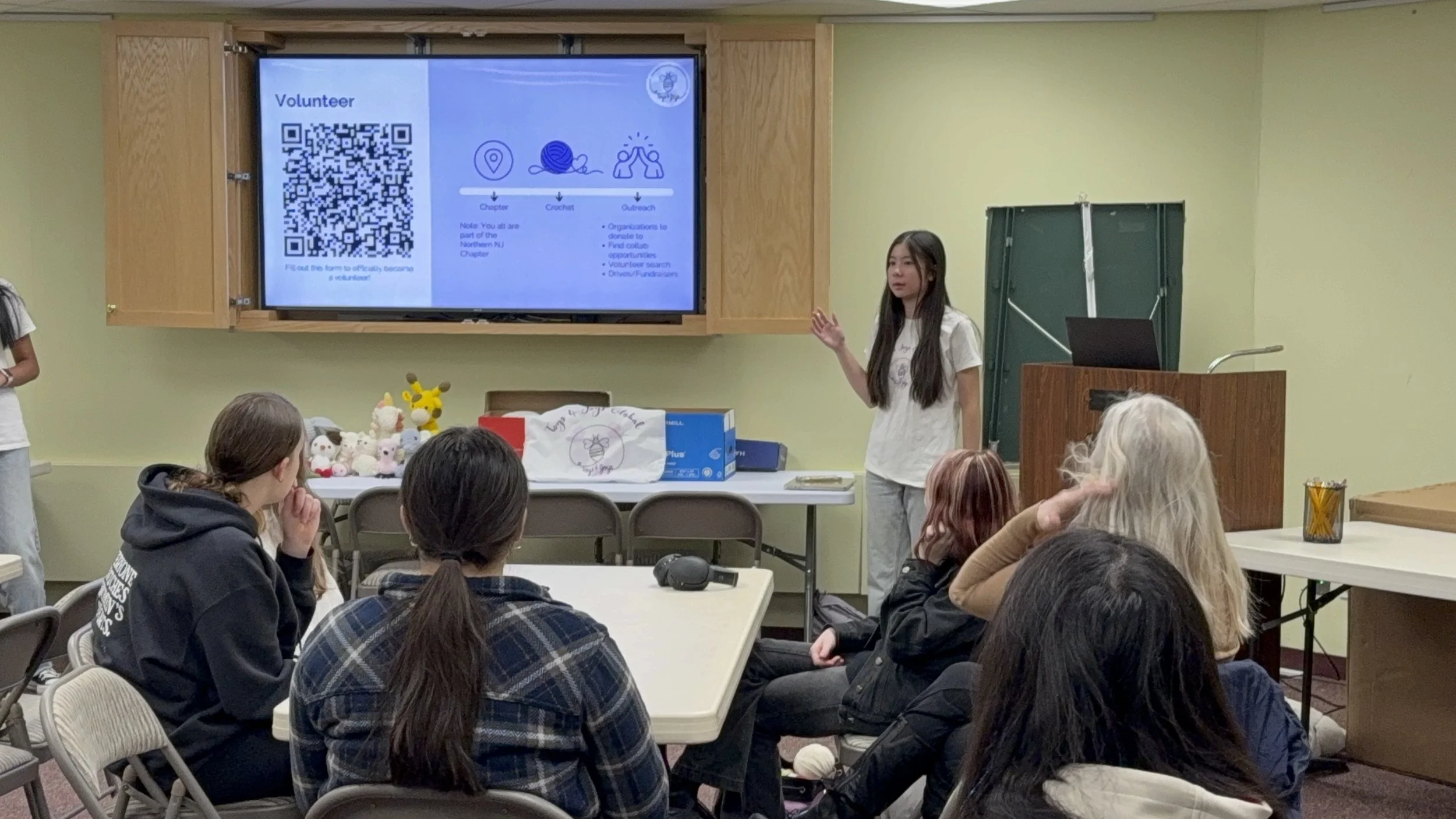 A young woman giving a presentation to a seated group of diverse people in a room with green walls. A large monitor displays information about volunteering, with a QR code on the left side.
