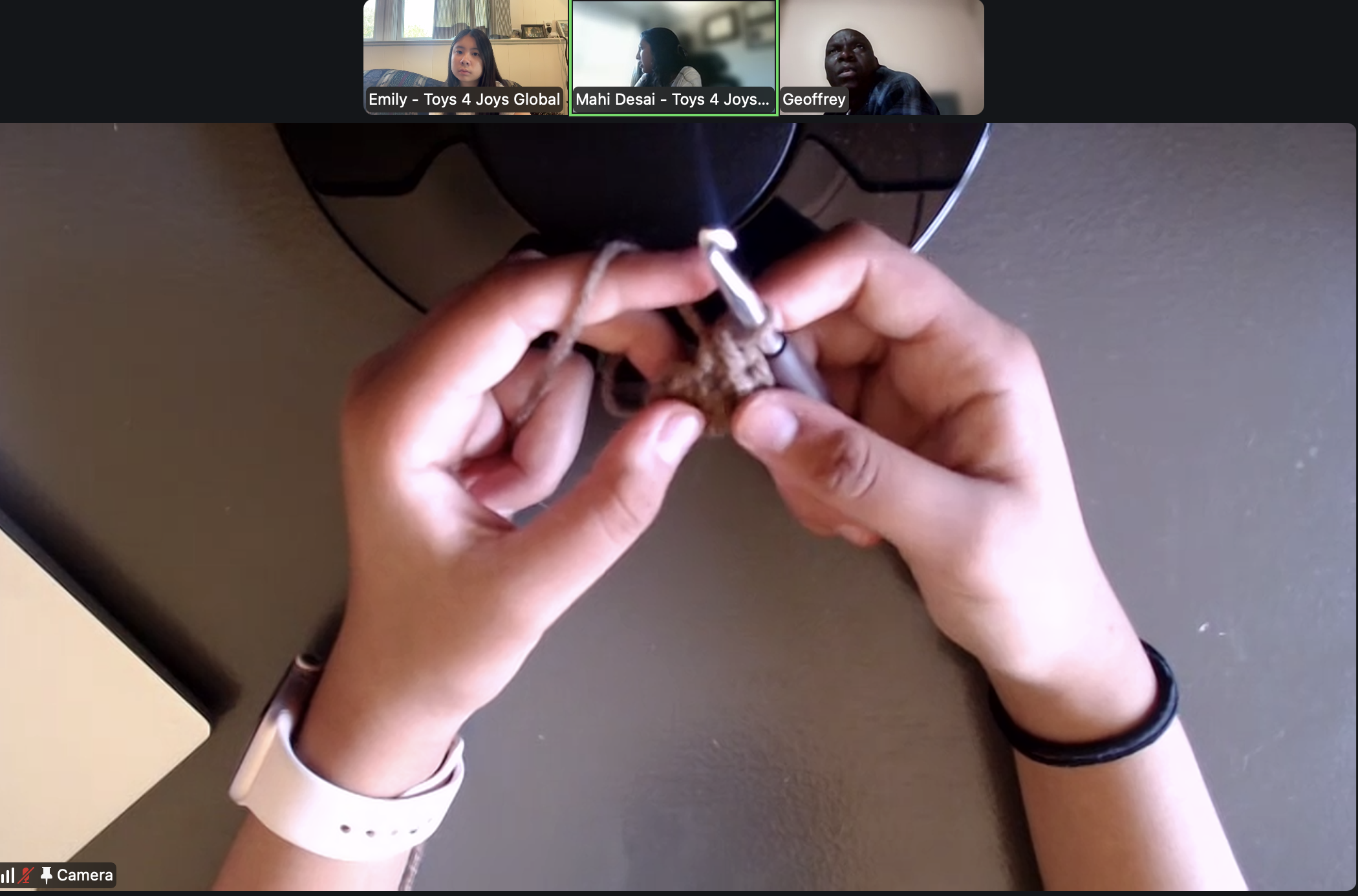Close-up of hands holding a crochet hook and yarn, working on a crochet project, on a gray surface, with a person wearing a black bracelet and a pink sleeve visible in the background. Video call interface showing three participants in small windows at the top.