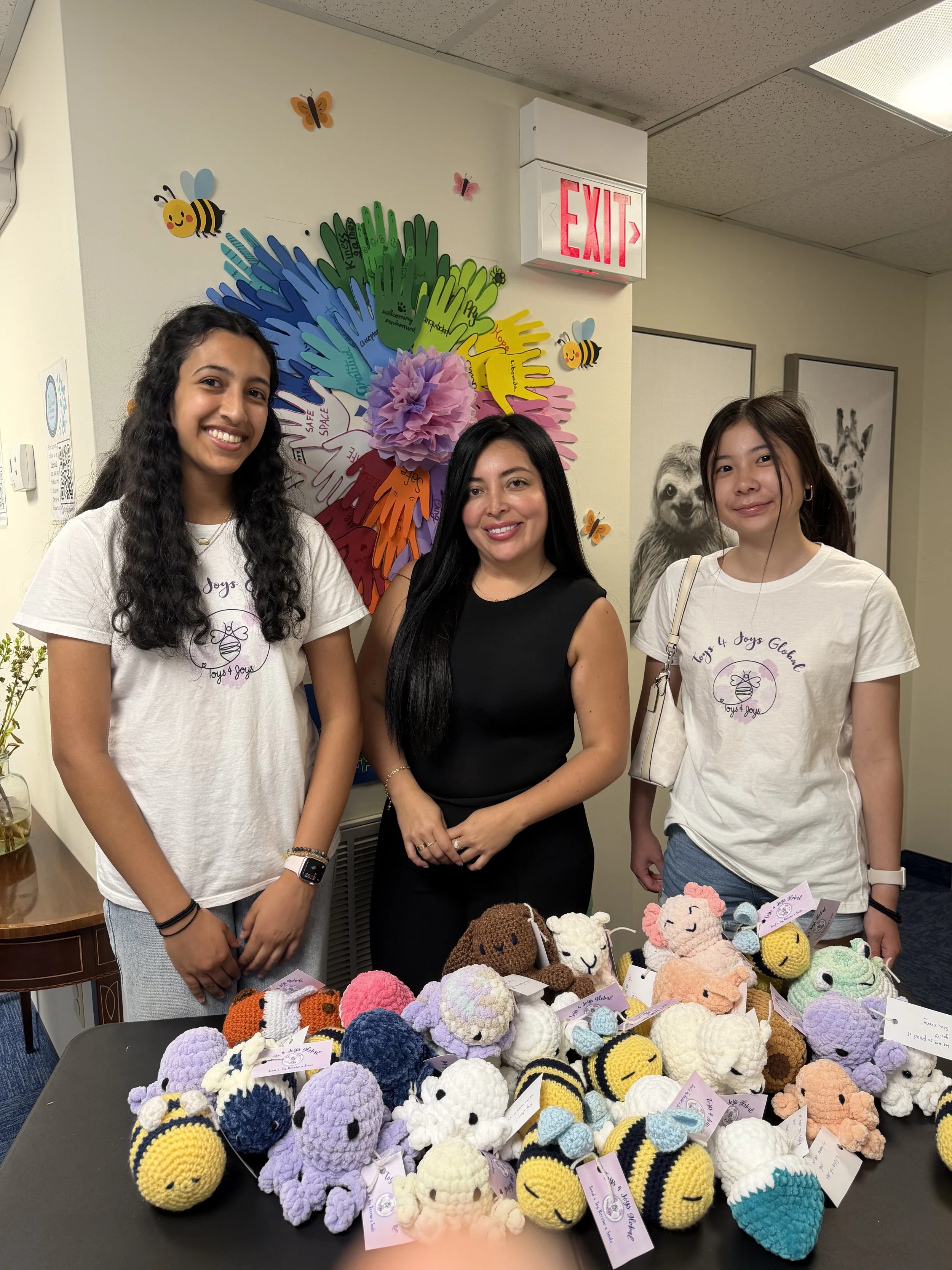 Three women standing behind a table with various colorful crocheted stuffed animals, with a large handmade wall decoration of handprints in different colors and bee and butterfly cutouts behind them.