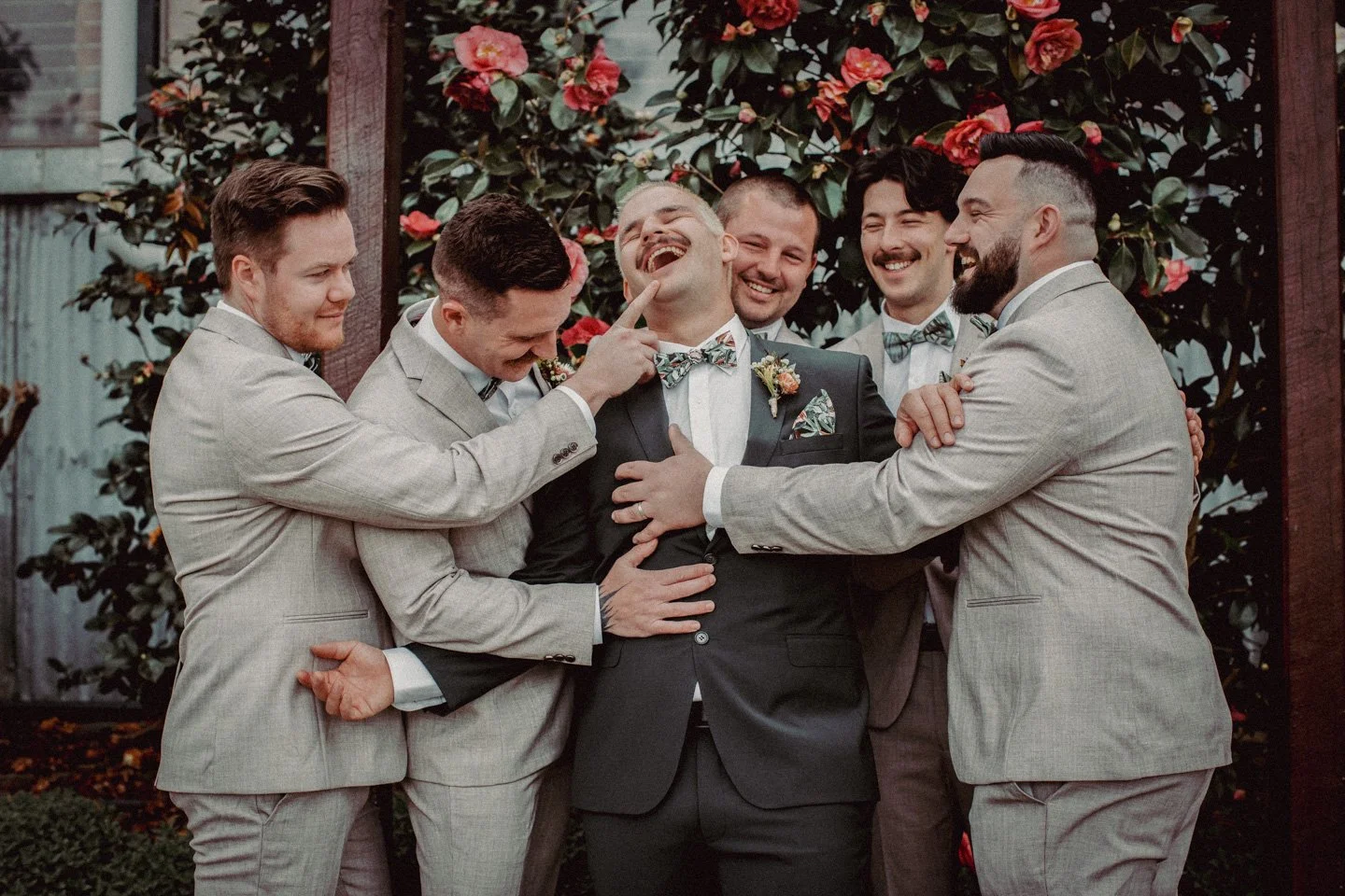 A groom and his friends are gathered together outdoors in front of blooming pink camellia flowers, sharing a joyful moment and laughter.