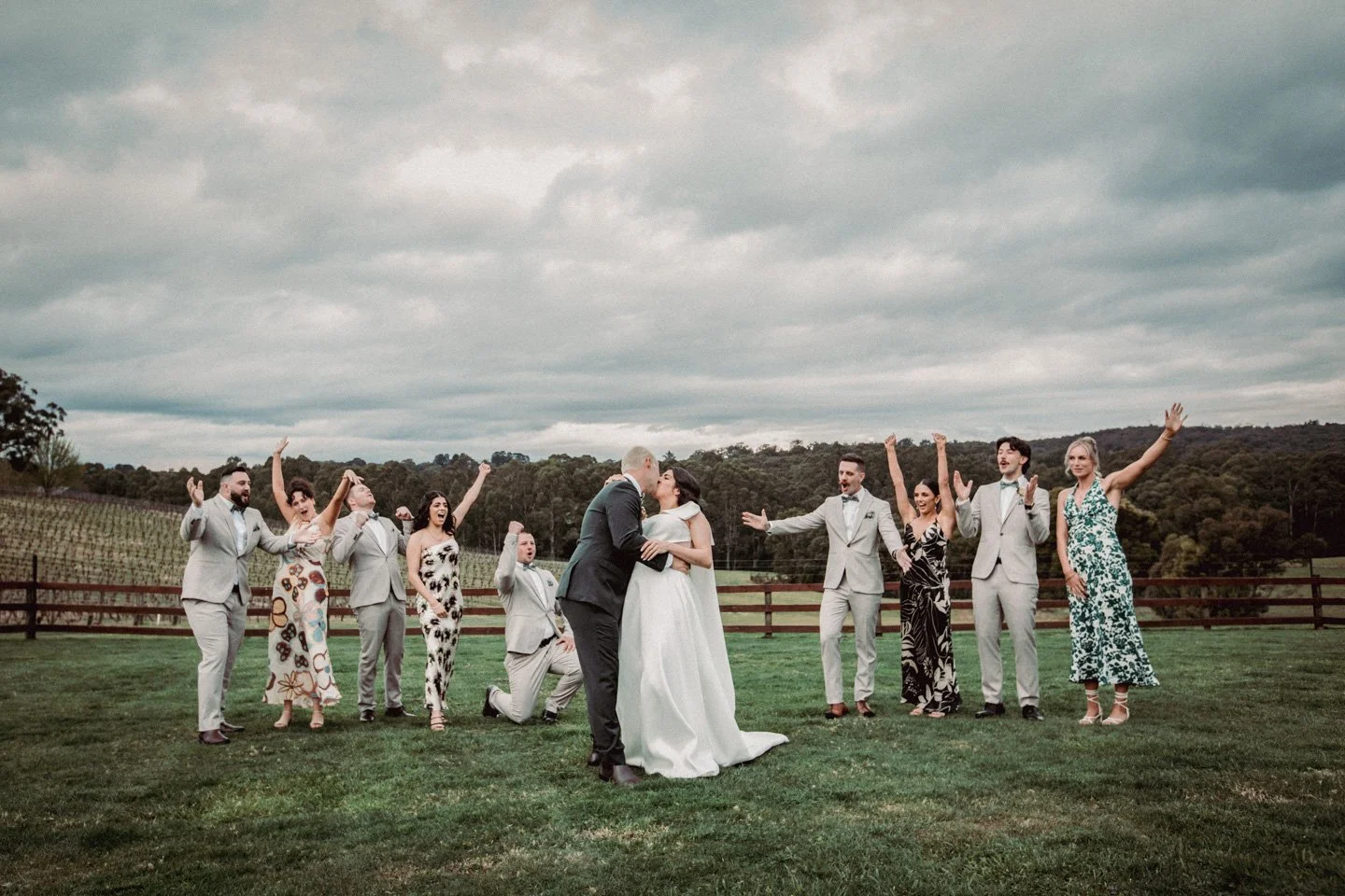 Wedding party standing on a grassy field outdoors, with a bride and groom kissing in the center, surrounded by guests celebrating with arms raised, against a cloudy sky and hilly landscape in the background at Yarra Ranges Estate.