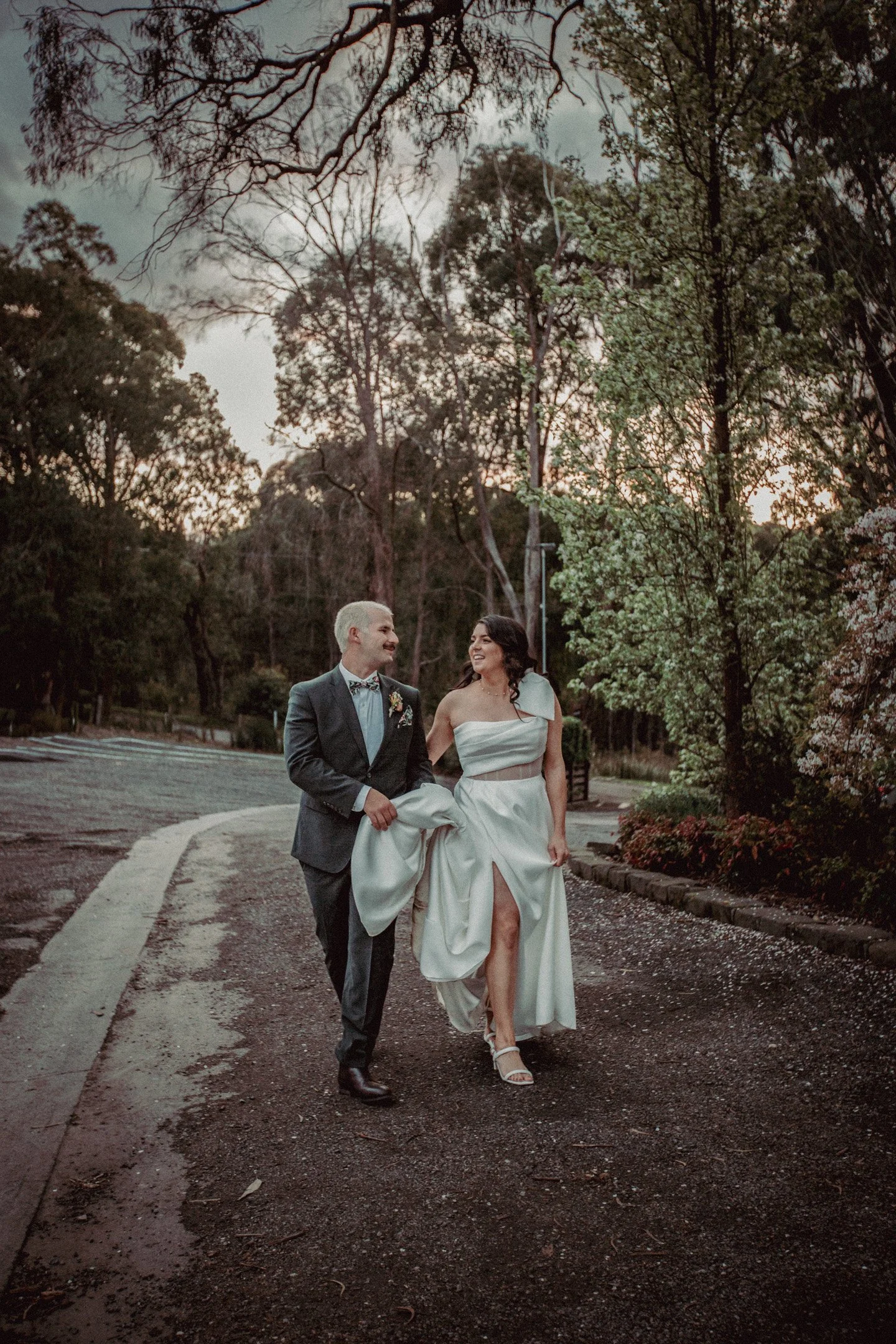 A newlywed couple walking outdoors on a dirt path surrounded by trees during sunset, with the groom in a dark suit and the bride in a strapless white wedding dress.