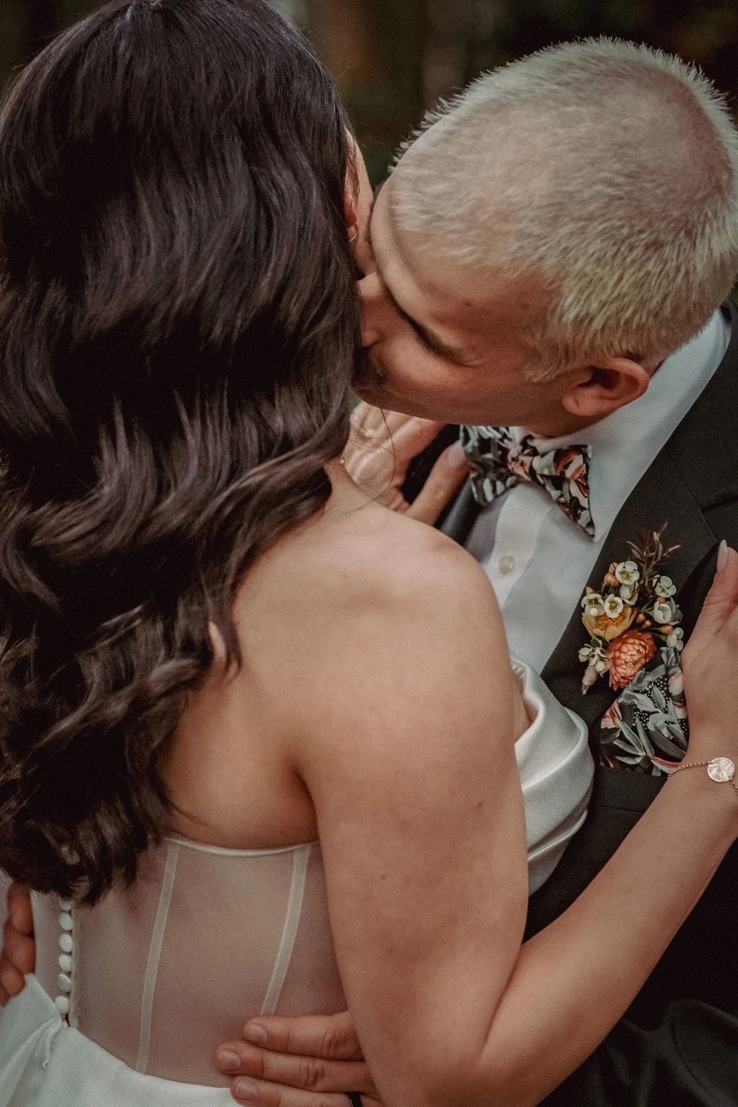 A couple, dressed in formal attire, engaged in a kiss, with the woman wearing a strapless dress with sheer details and the man in a suit with a floral bow tie and matching boutonniere.
