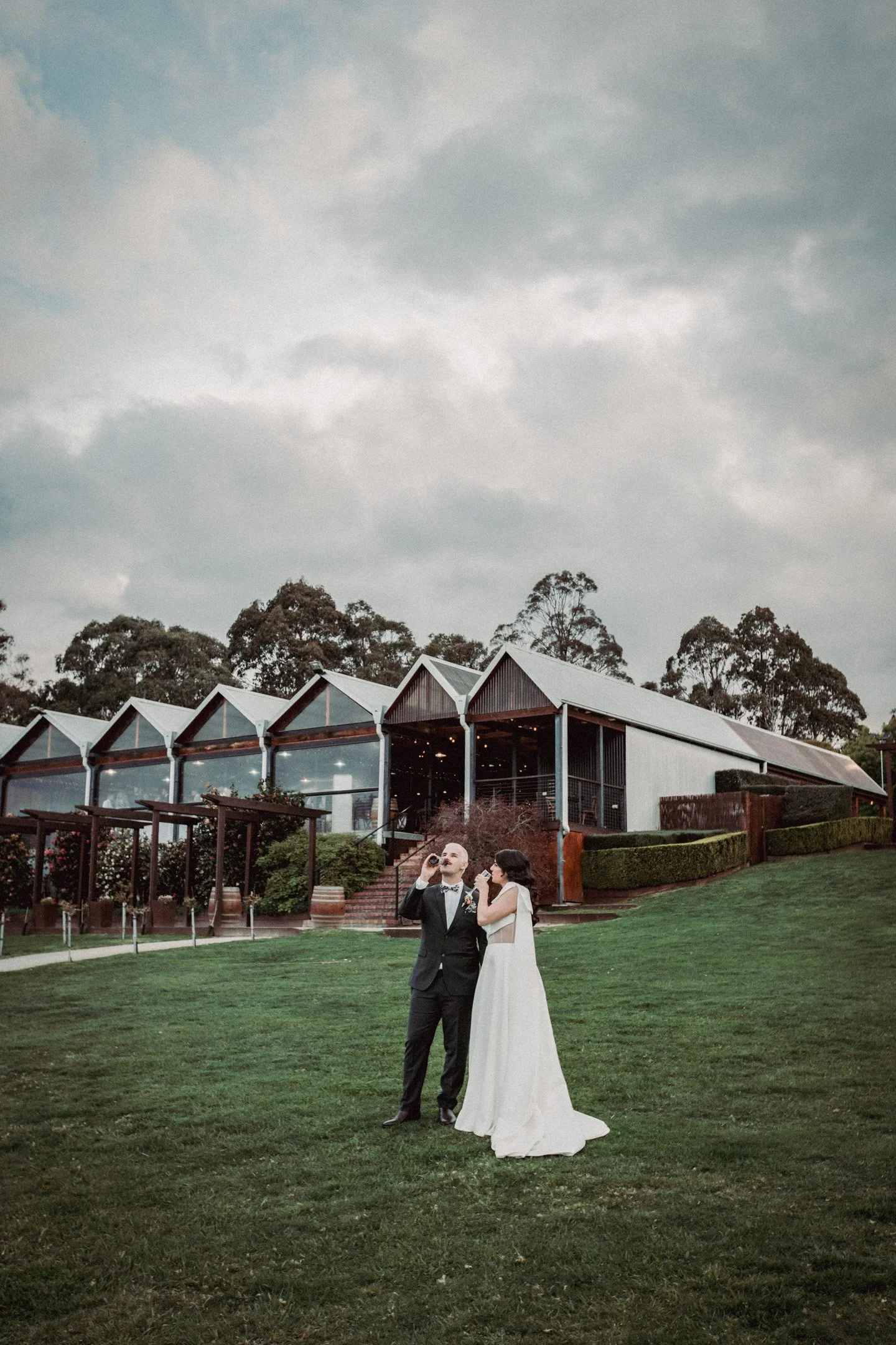 A bride and groom standing on a grassy area outside Yarra Ranges Estate, with trees in the background and cloudy sky above, during a wedding celebration.
