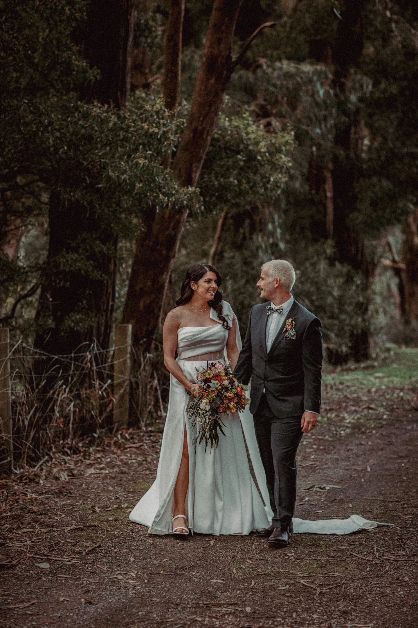 A newlywed couple walking hand in hand on a woodland trail, with the bride holding a bouquet of flowers and wearing a white dress, and the groom in a dark suit with a bow tie, surrounded by tall trees.