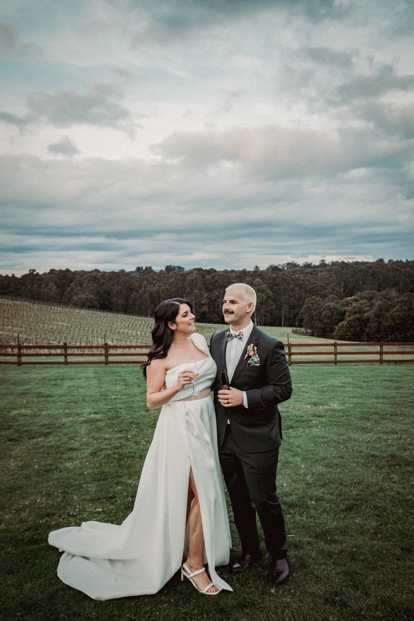 A bride and groom standing together outdoors on their wedding day, holding drinks and smiling at each other, with a vineyard and cloudy sky in the background.