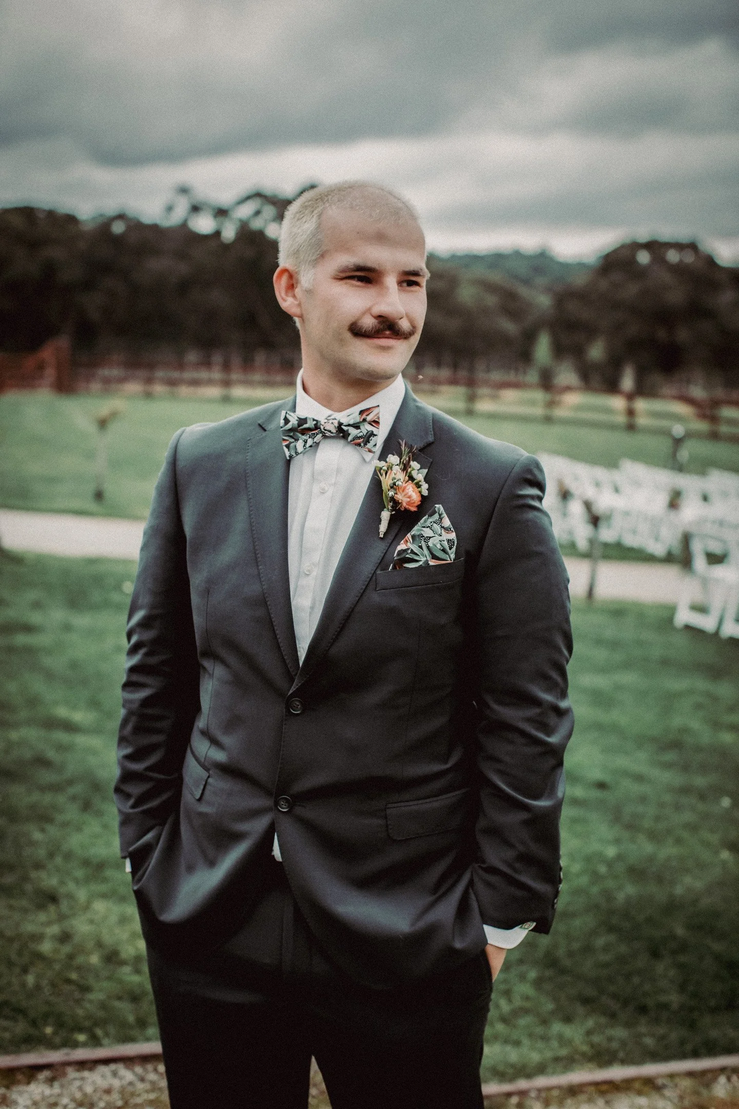 A groom in a dark suit with a colourful bow tie and floral accessories standing outdoors on a grassy field with cloudy sky in the background at Yarra Ranges Estate.