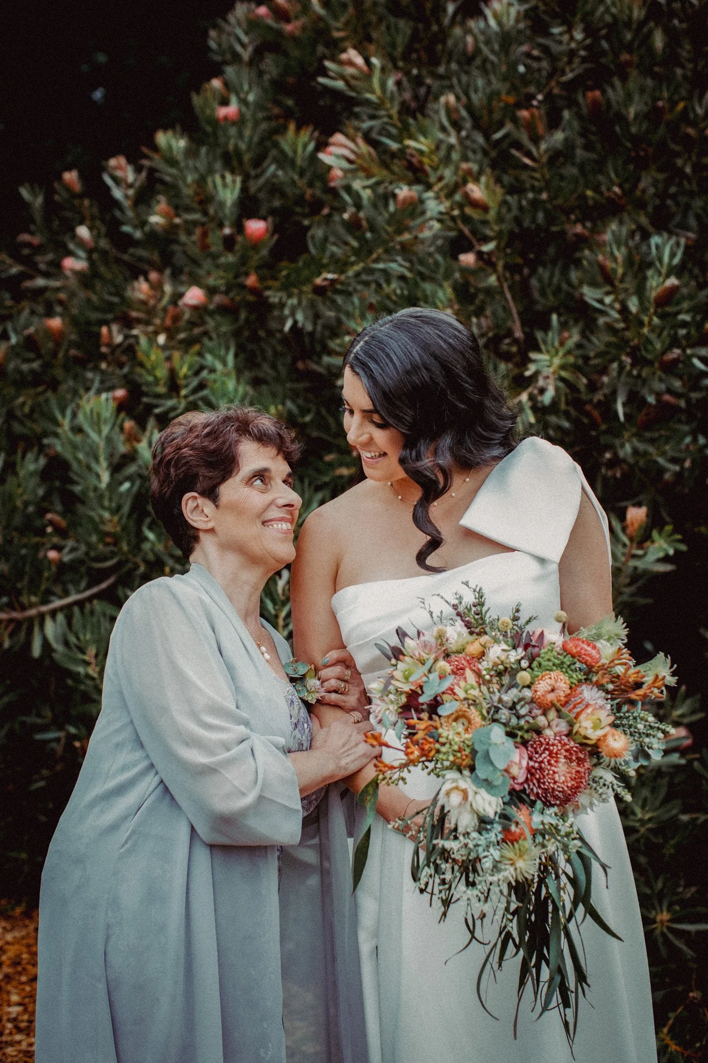 A bride holding a large bouquet of colorful flowers and her mother, with a background of green foliage.