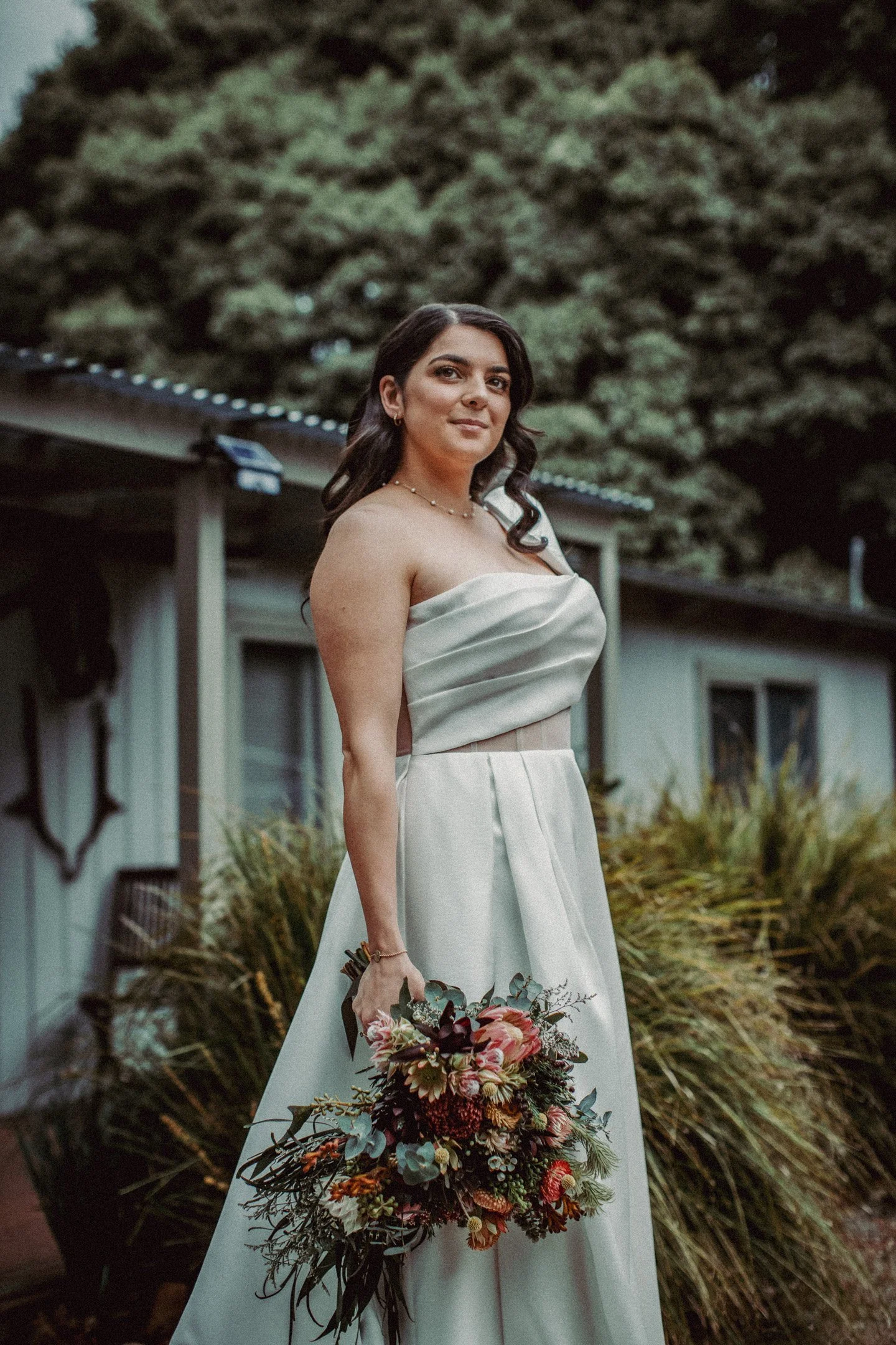 Bride in white wedding dress holding a bouquet standing outdoors in front of green foliage and a building.