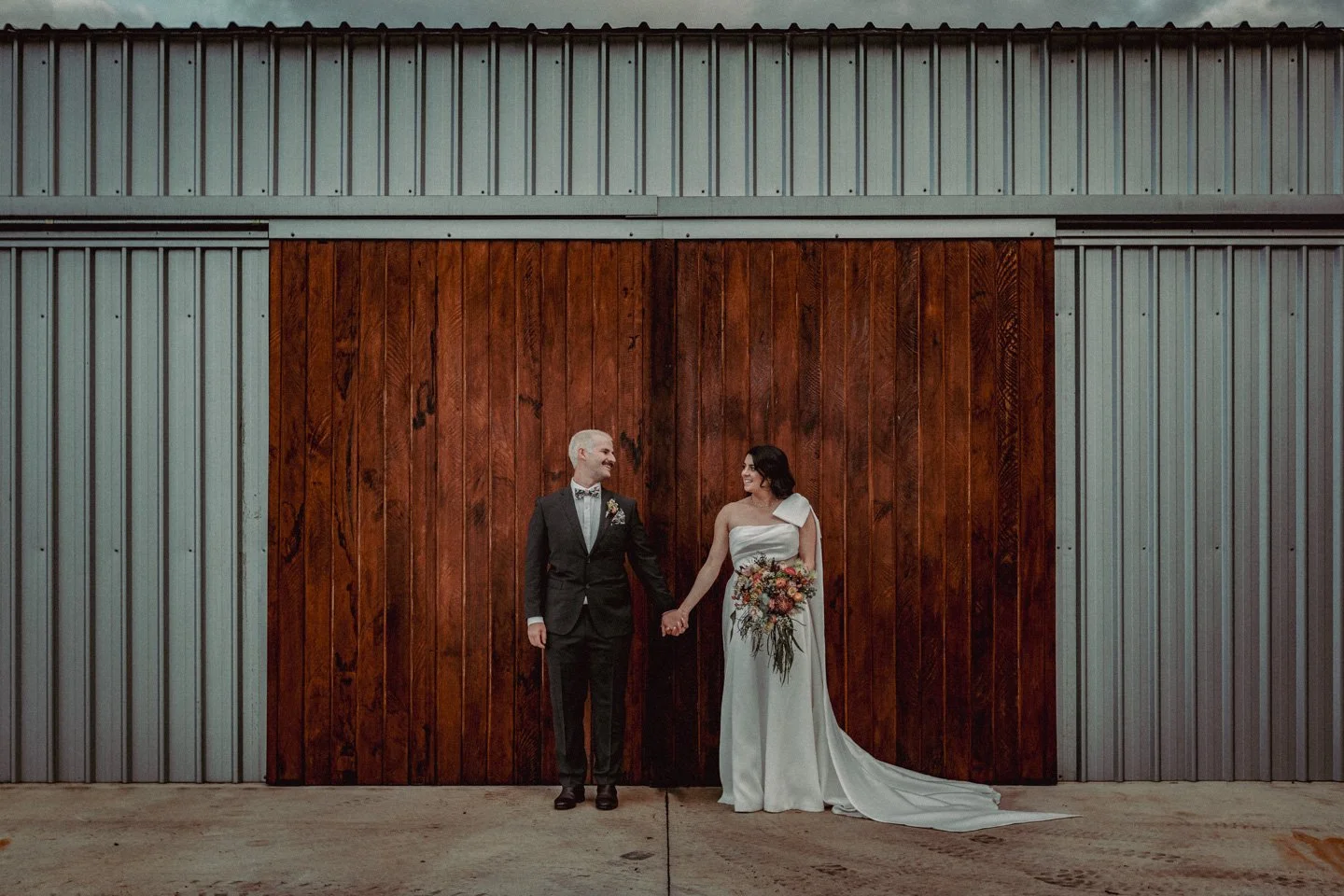 A bride and groom holding hands and smiling at each other in front of a wooden barn door with metal siding at Yarra Ranges Estate.