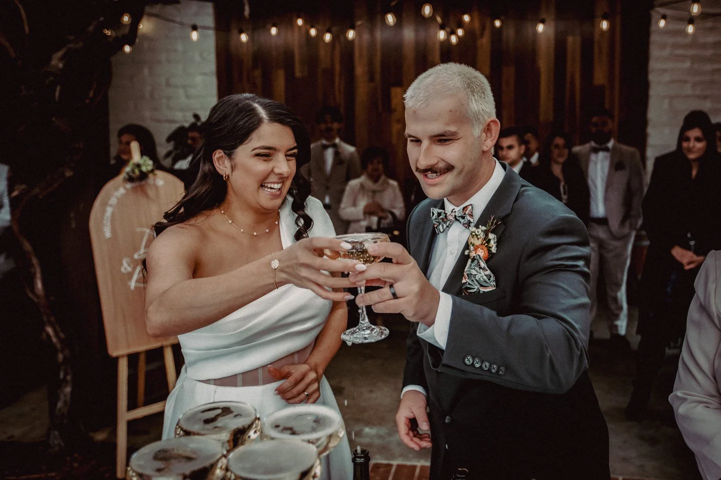 A wedding celebration with a bride and groom toasting with drinks, surrounded by guests in a warmly decorated indoor area at Yarra Ranges Estate.
