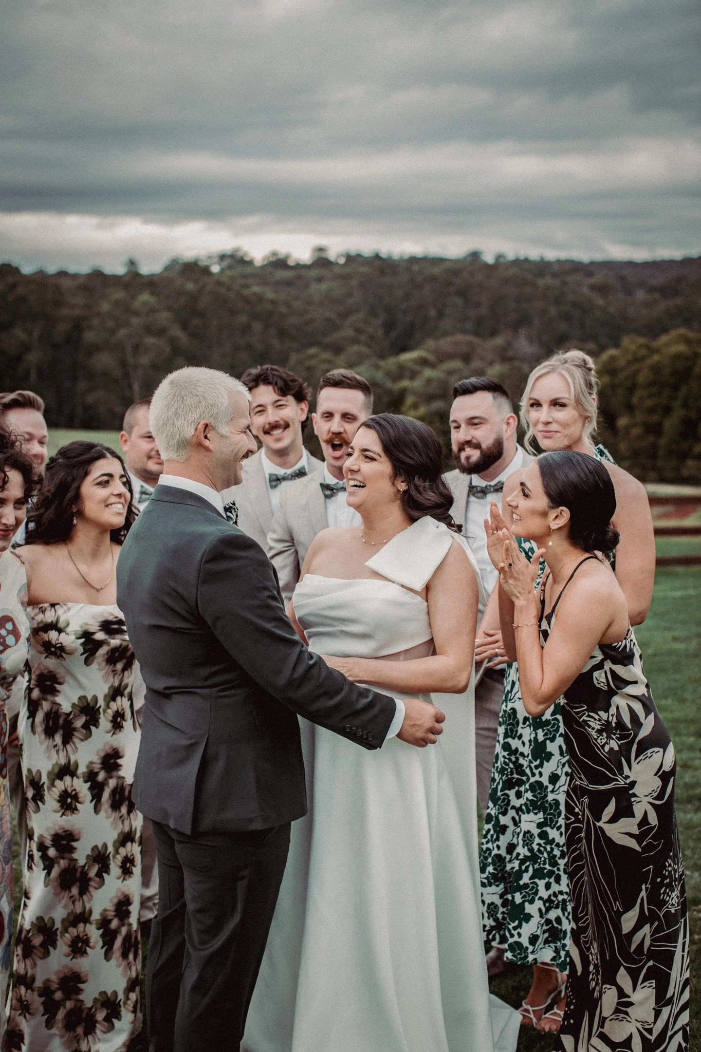 A bride and groom exchange vows outdoors among smiling guests on a cloudy day, with a green landscape background.