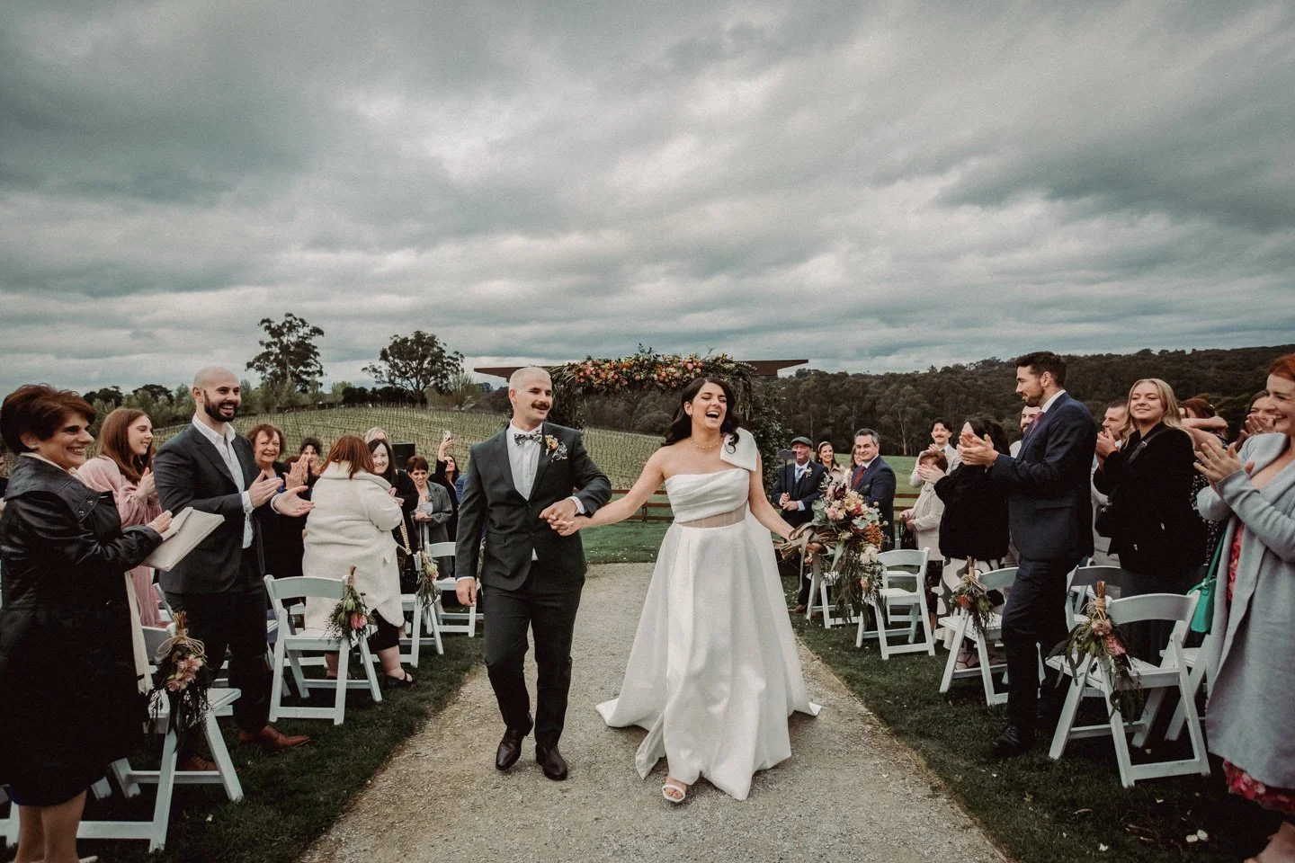 A newlywed couple walking down the aisle at their outdoor wedding ceremony, surrounded by smiling guests clapping, on a cloudy day in a scenic Yarra Ranges Estate.