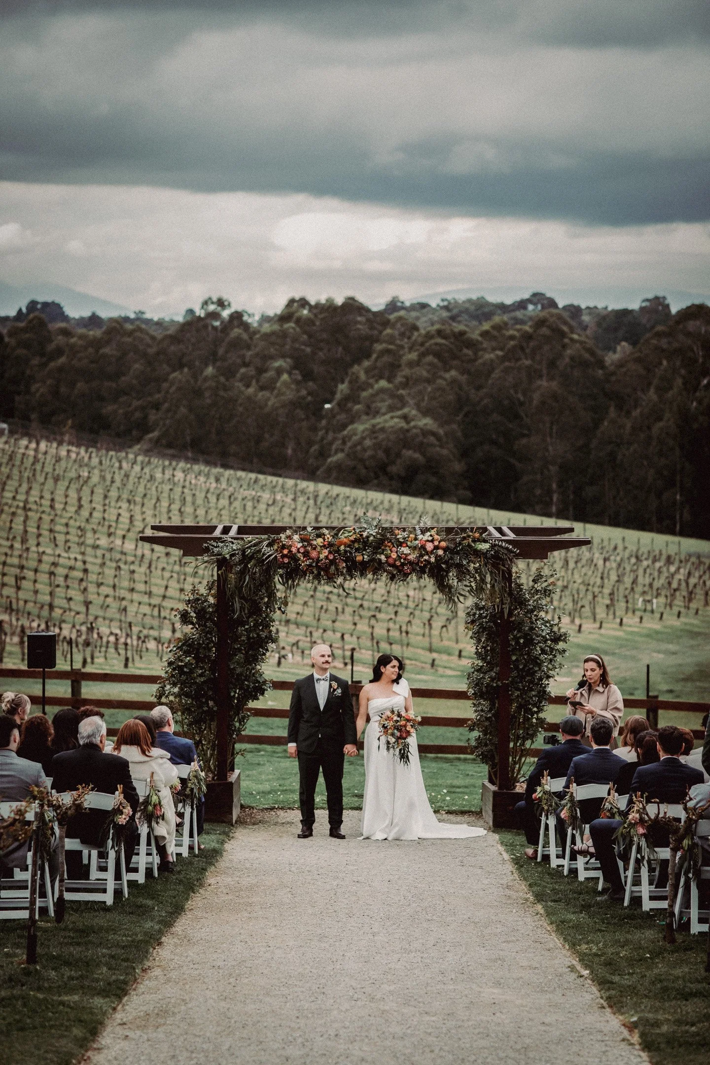 A bride and groom holding hands under an outdoor wedding arch decorated with flowers, with guests seated on either side and a vineyard in the background, at Yarra Ranges Estate.