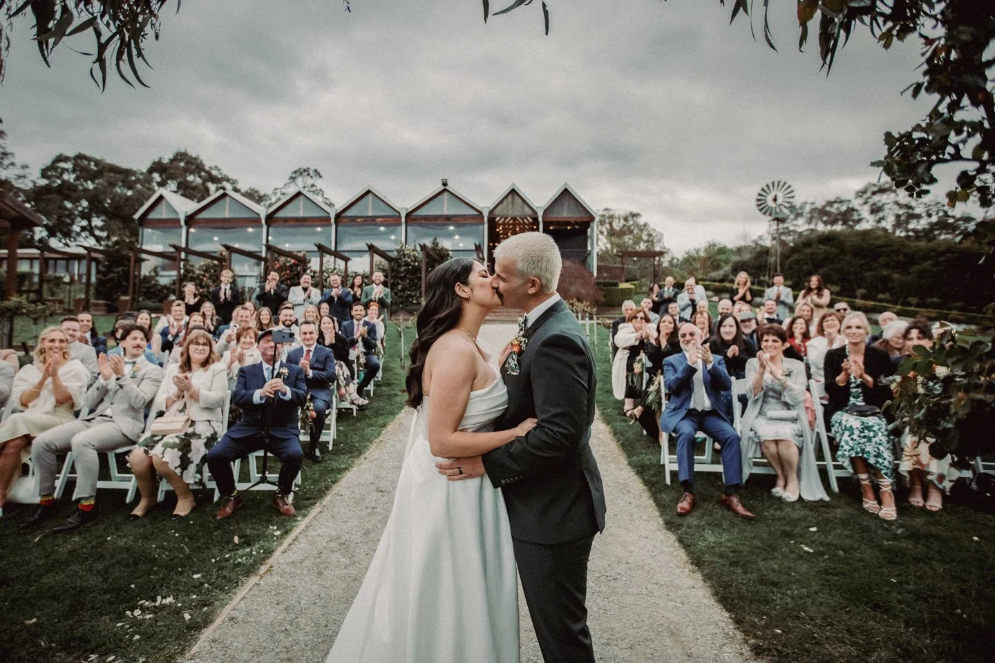 Bride and groom kissing during outdoor wedding ceremony with seated guests clapping in the background at at Yarra Ranges Estate.