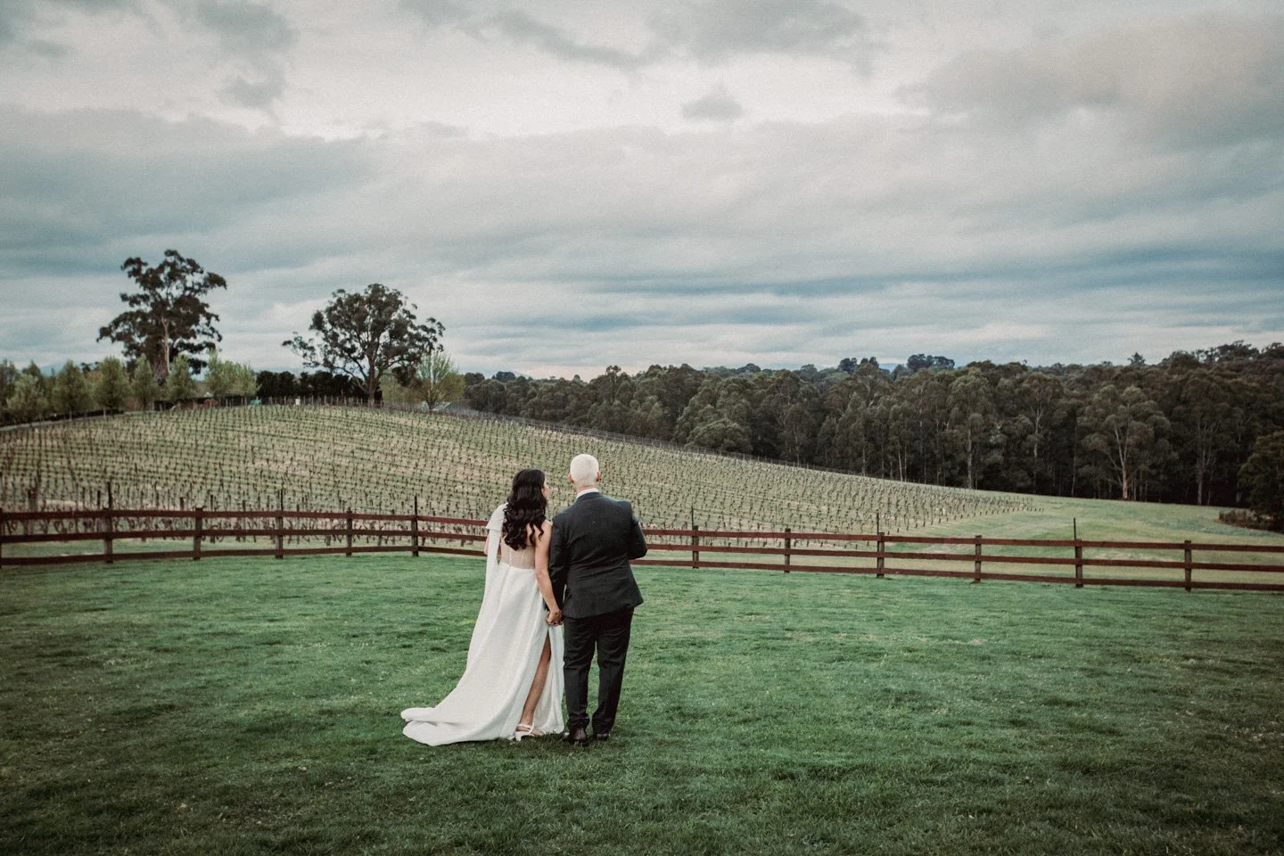 Bride and groom holding hands, walking on a lush green lawn towards a vineyard in a rural landscape under a cloudy sky.