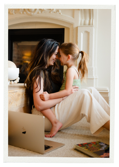 A woman and a young girl, possibly mother and daughter, sitting on the floor in front of a fireplace, touching foreheads and smiling at each other. A laptop and a colorful notebook are nearby.