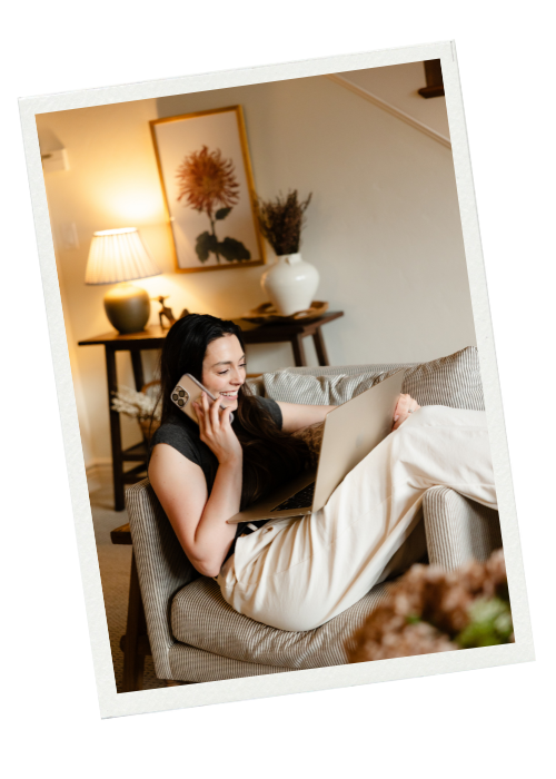 A young woman sitting on a beige armchair, talking on a cellphone, with a laptop on her lap in a cozy living room with warm lighting, a small table with a lamp, framed artwork, and decorative vases in the background.