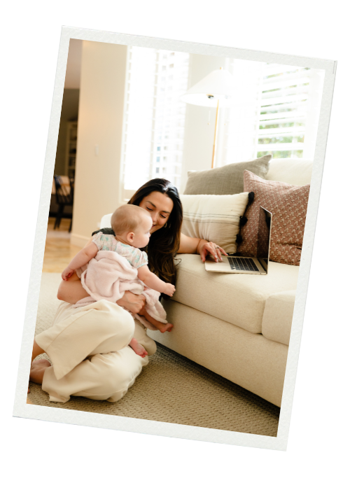 A woman sitting on the floor with a baby in her lap, using a laptop on a couch in a bright living room with large windows.