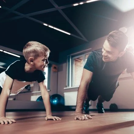 A father and young son doing push-ups together in a gym or fitness room with exercise equipment in the background.