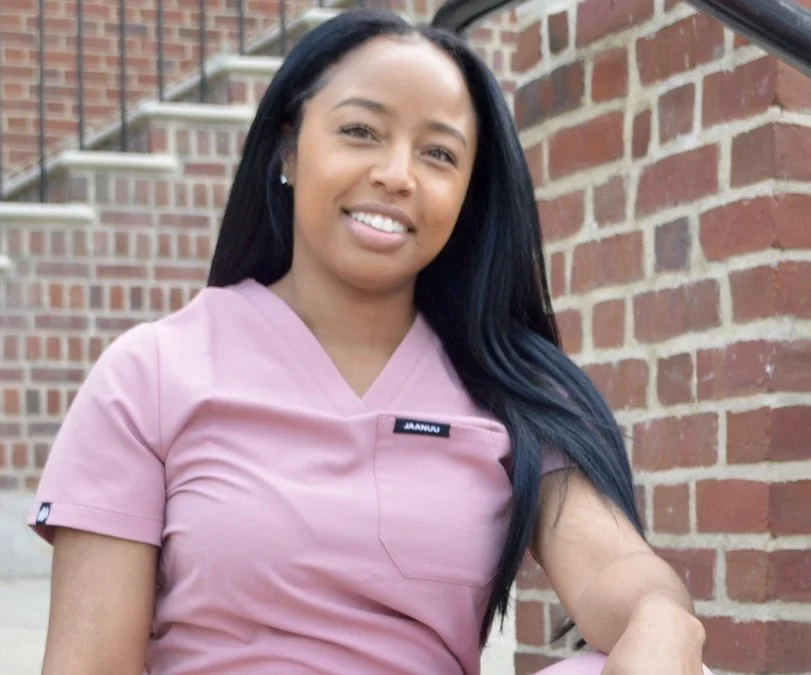 Young woman in pink scrubs sitting on outdoor steps next to a brick wall.