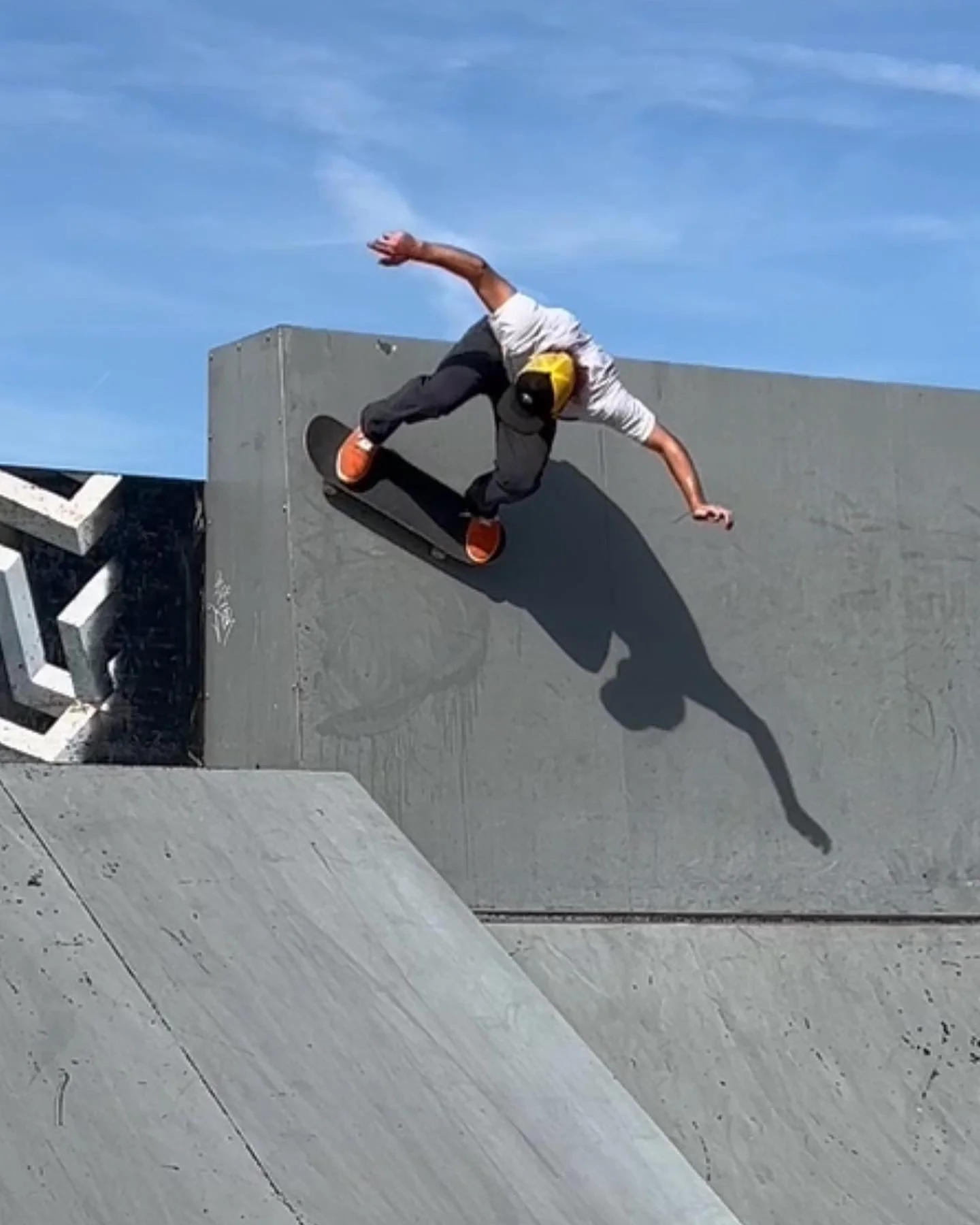 A skateboarder wearing a yellow hat, white t-shirt, and dark pants performing a trick on the side of a skate ramp at an outdoor skate park under a blue sky.