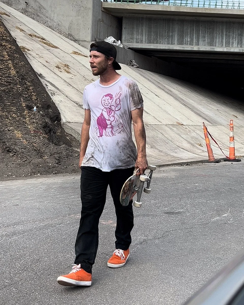 A man walking on a street, holding a skateboard, wearing an orange pair of shoes, black pants, and a white T-shirt with a pink cartoon graphic. The man is wearing a black backwards cap, and has dirt stains on his shirt. The background shows an underpass with orange traffic cones and dirt on the slope.