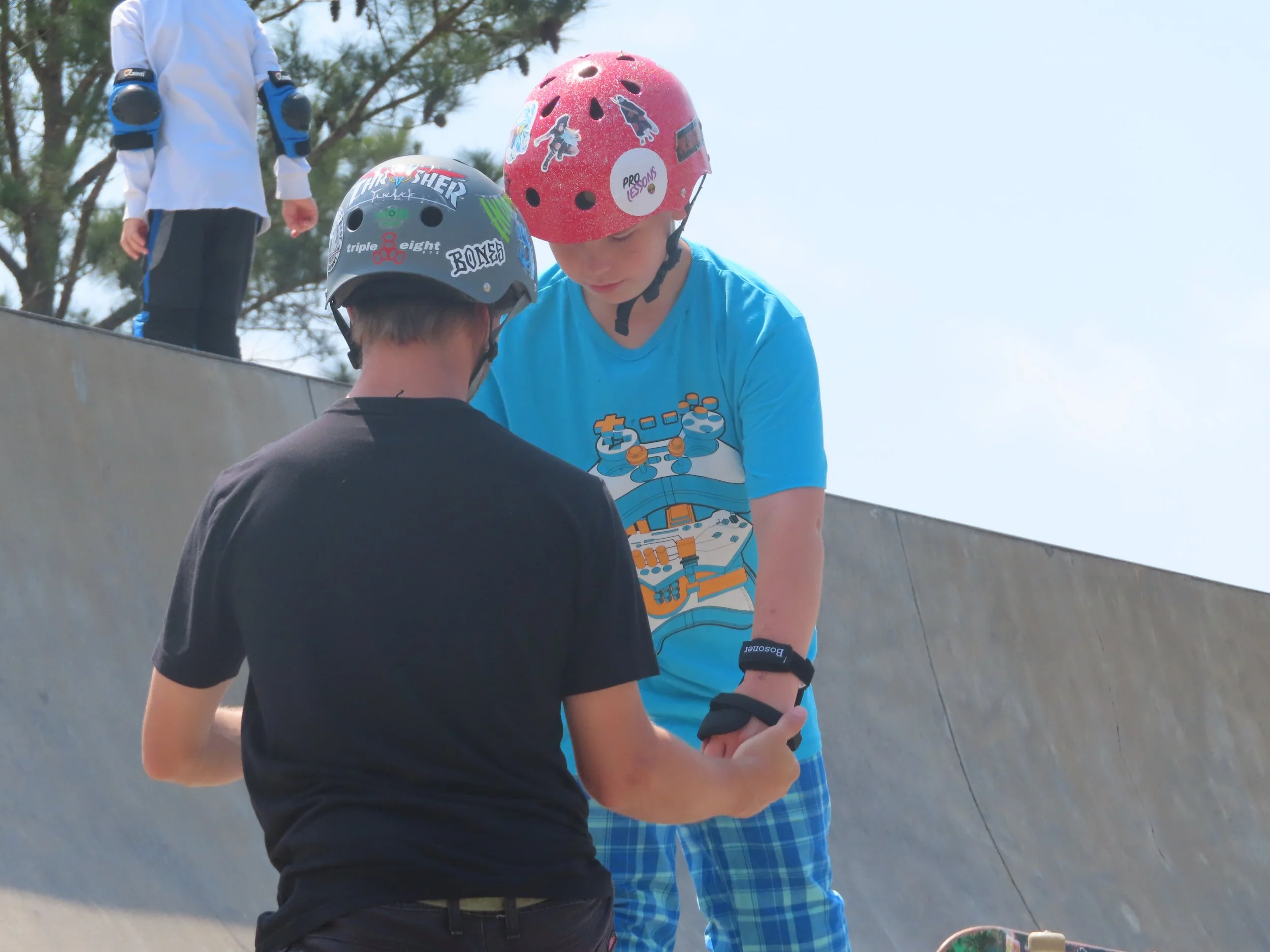 Two boys wearing helmets at a skate park, one in a black shirt and the other in a blue shirt, with an adult in the background.
