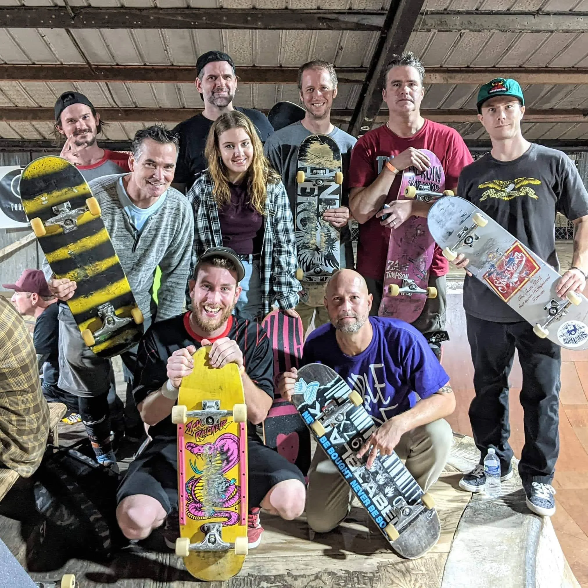 A group of ten people holding skateboards, posing indoors at a skatepark.