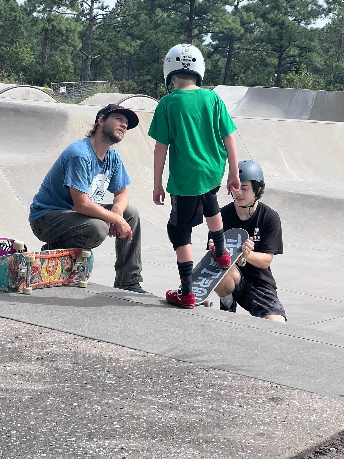 A young boy with a helmet is receiving skateboarding instructions from two men, one in a blue shirt and another kneeling with a helmet, at a skatepark. The boy is standing on a skateboard while the others assist him.
