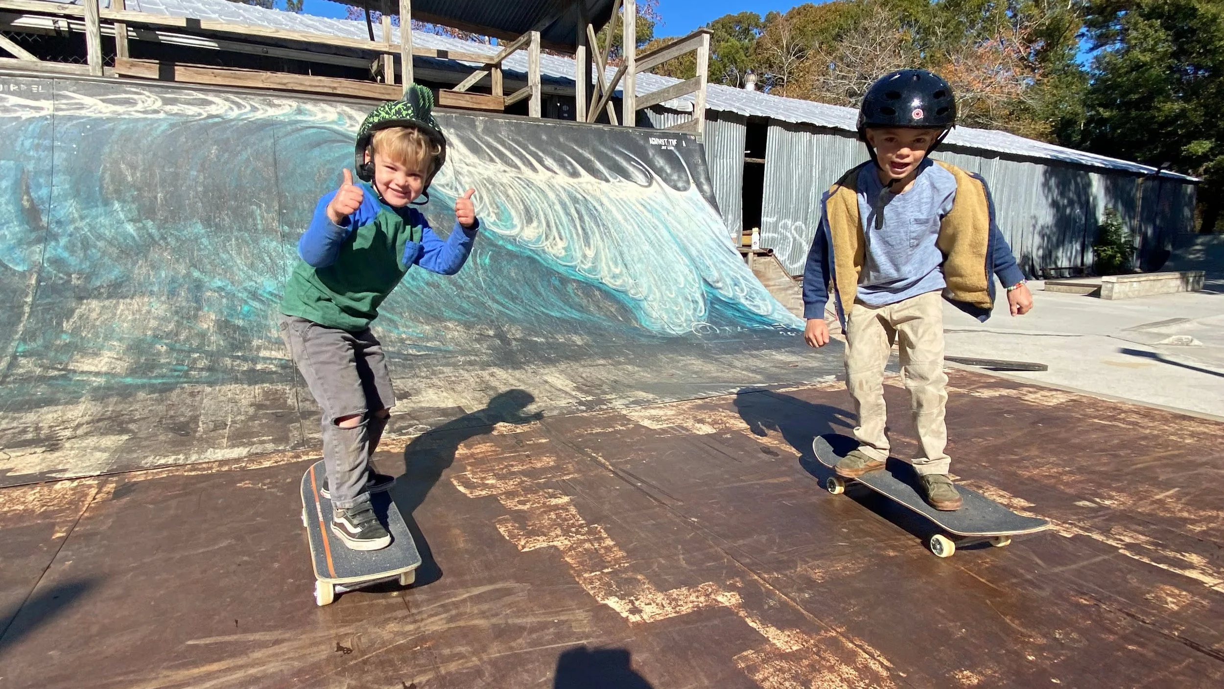 Two young boys skateboarding at an outdoor skate park, with one giving a thumbs-up and smiling, the other wearing a helmet. A painted ramp with ocean waves is in the background.