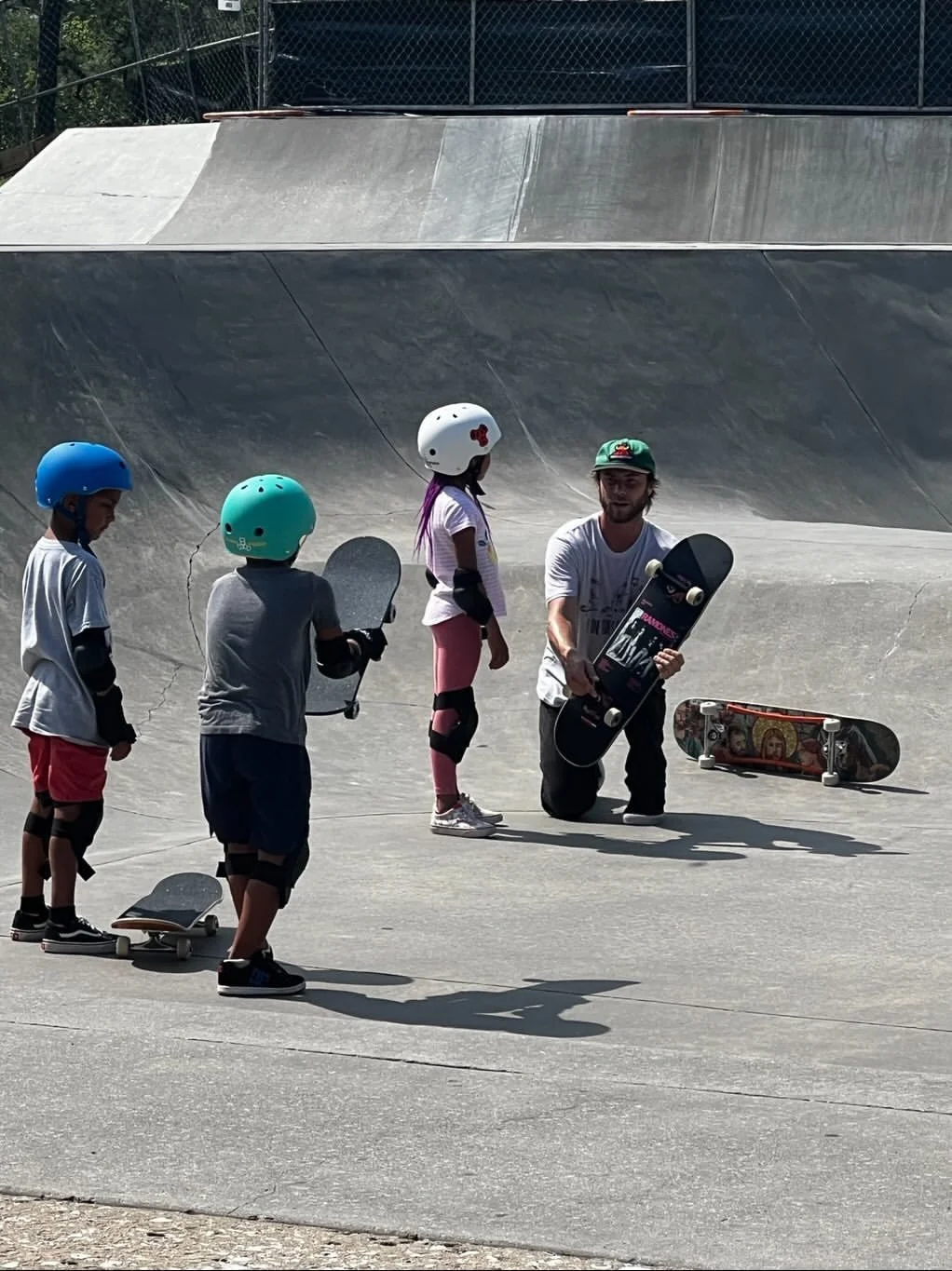 A skate instructor kneeling on the concrete skatepark with three children, all wearing helmets, holding skateboards during a lesson.