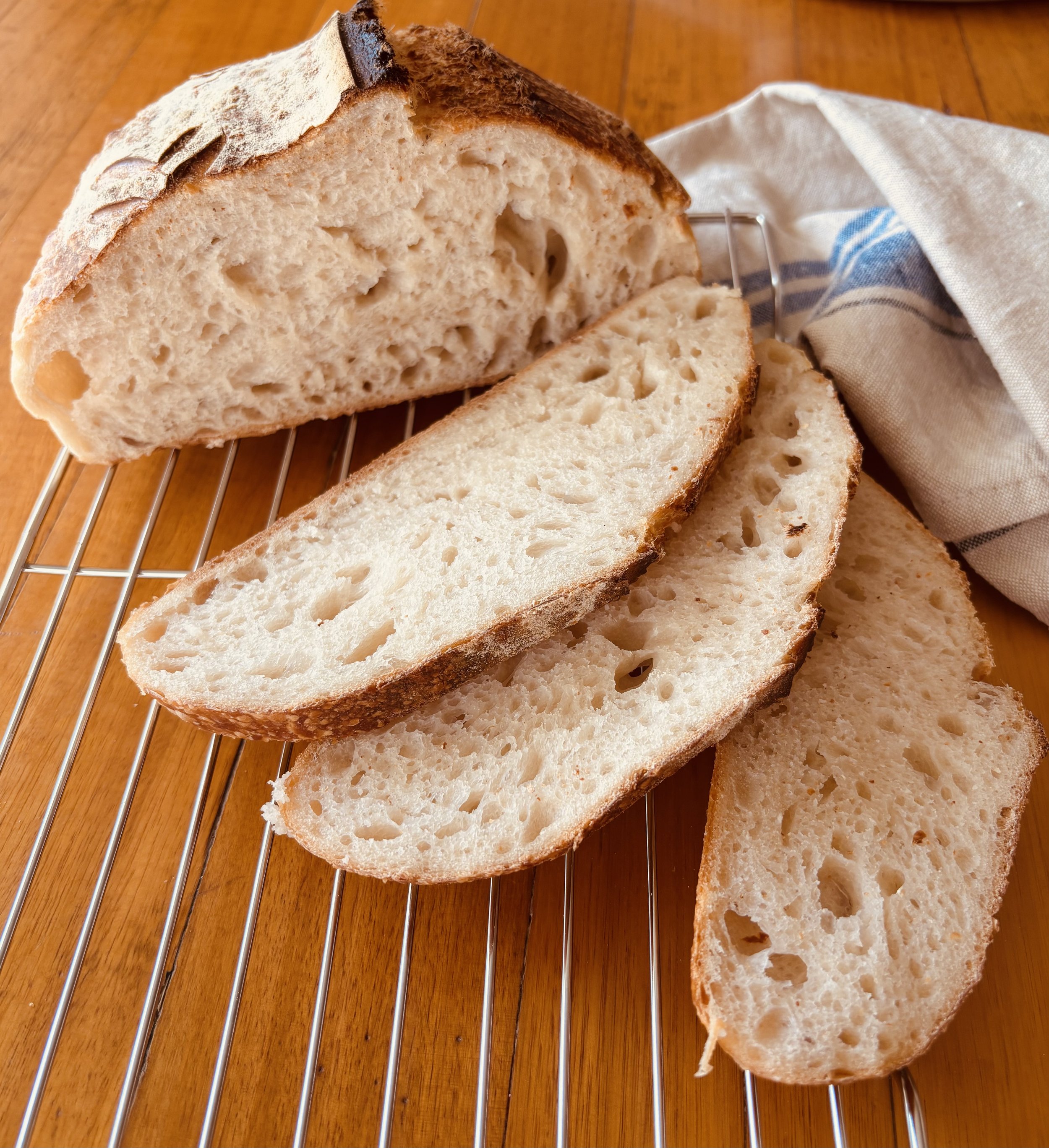 Sliced loaf of rustic bread on a cooling rack, next to a white cloth on a wooden surface.