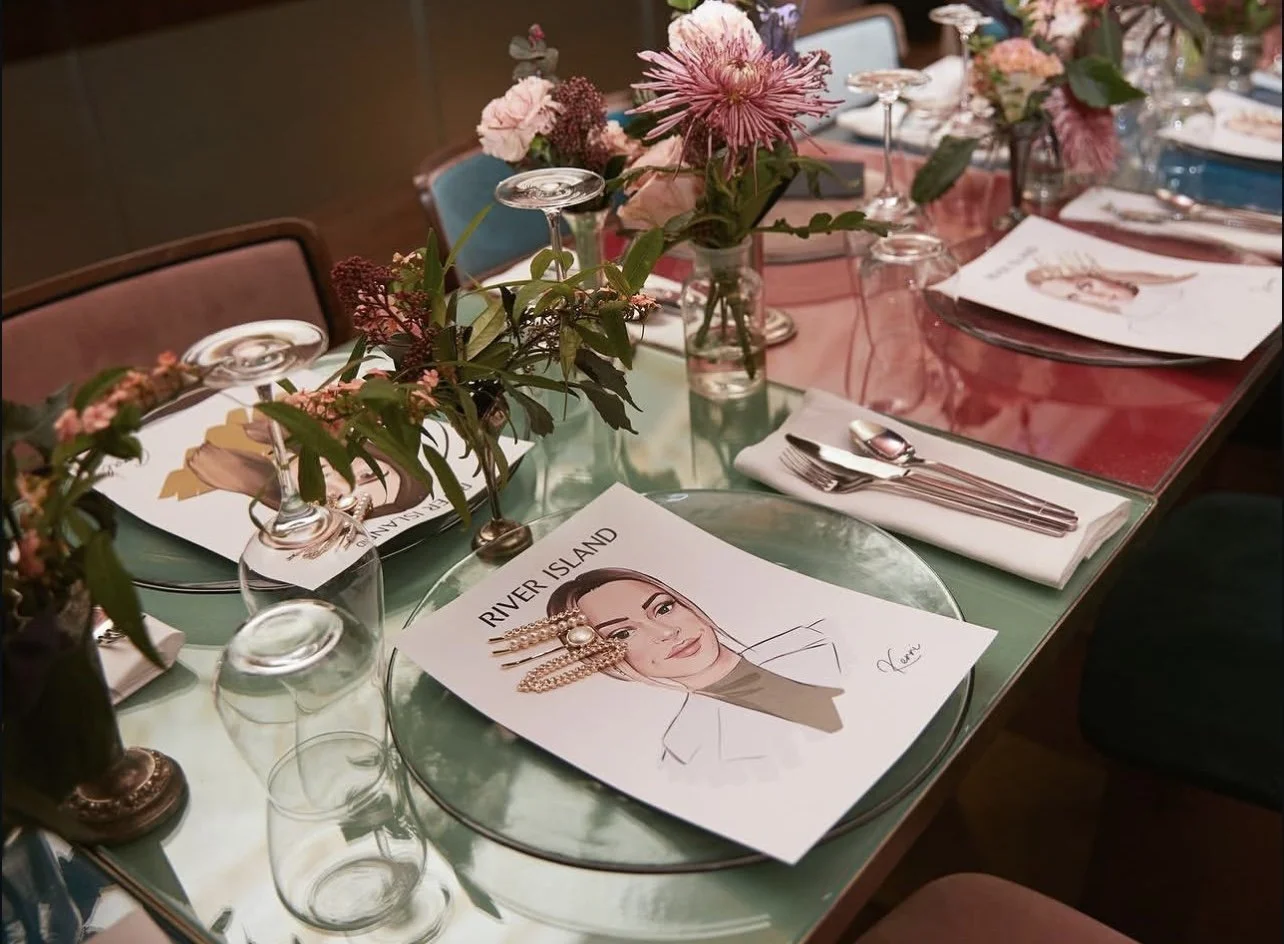 A table set for a formal event with floral centerpieces, wine glasses, and printed menus. The menus feature a drawing of a woman with the title "River Island."