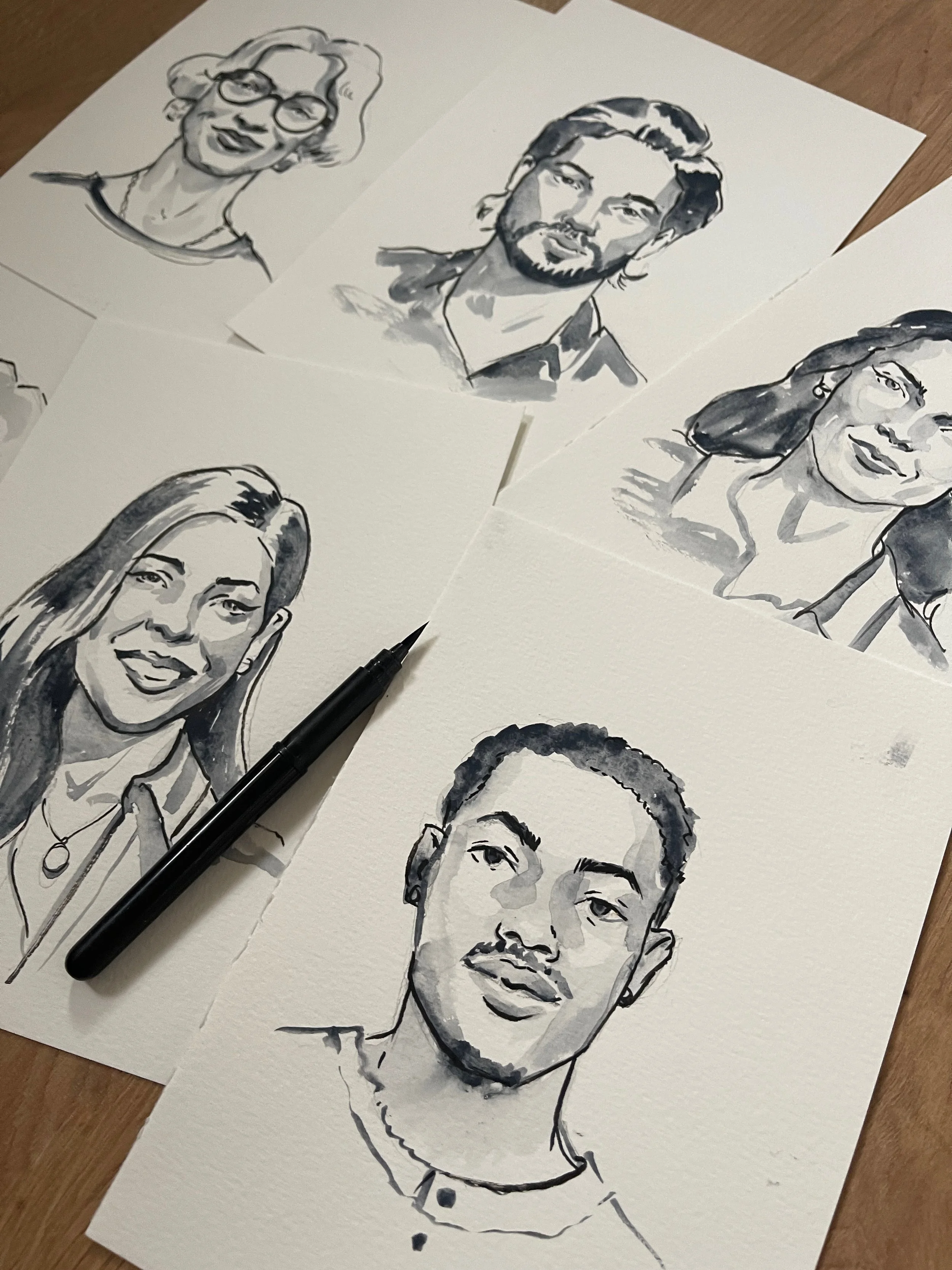 Collection of black and white watercolor portraits of diverse people on paper sheets, with a black pen resting on one of the sheets.