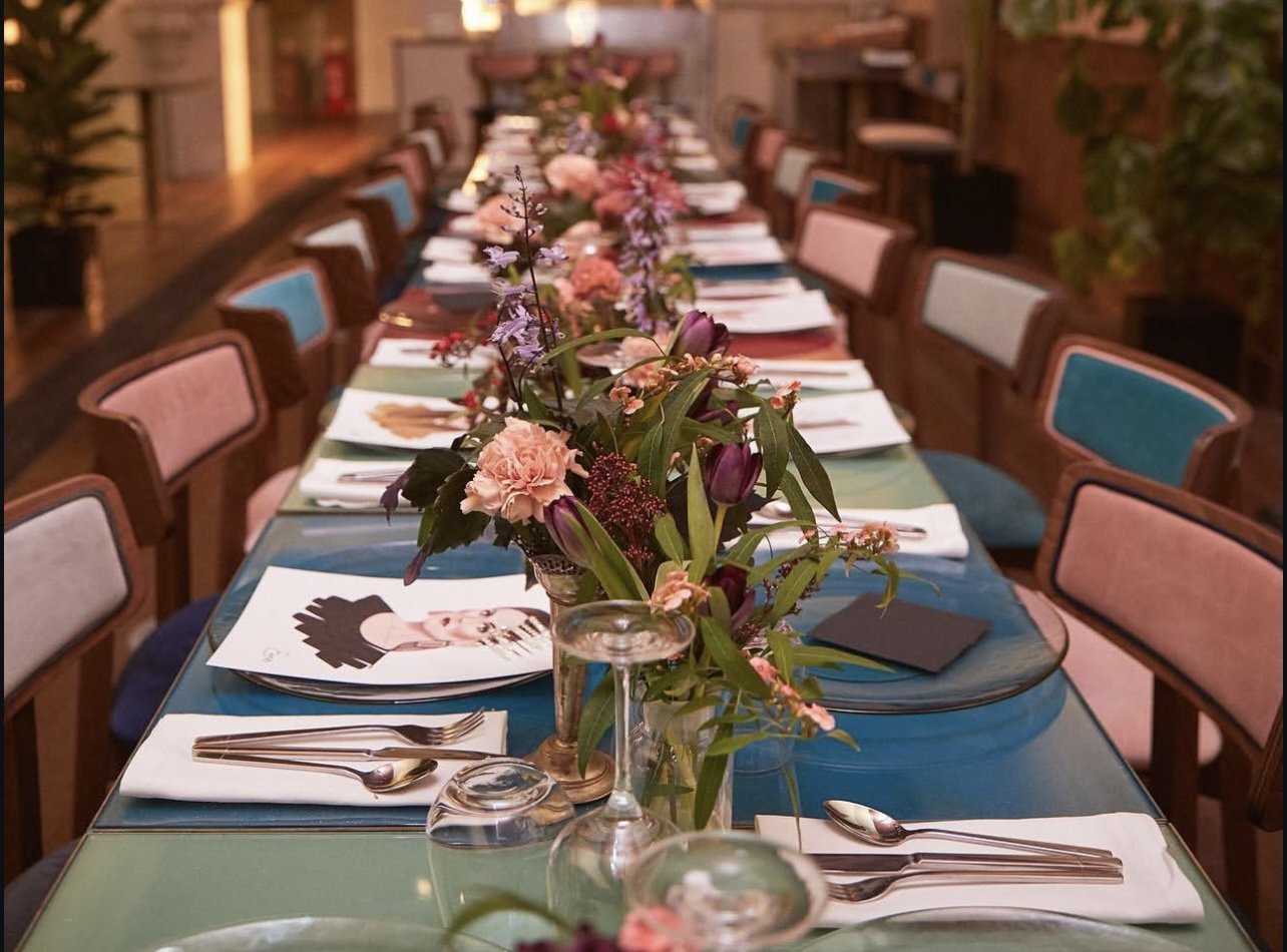 Long dining table decorated with pink and purple flowers in vases, set with white napkins, silverware, and black menus in a warmly lit restaurant or event space.