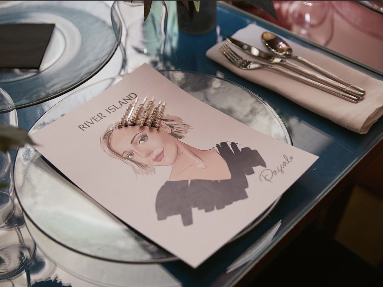 Table setting with a pamphlet titled 'River Island' featuring a woman's illustrated portrait and five sparkly hair clips on top, silverware on a napkin, and a glass plate on a glass table.