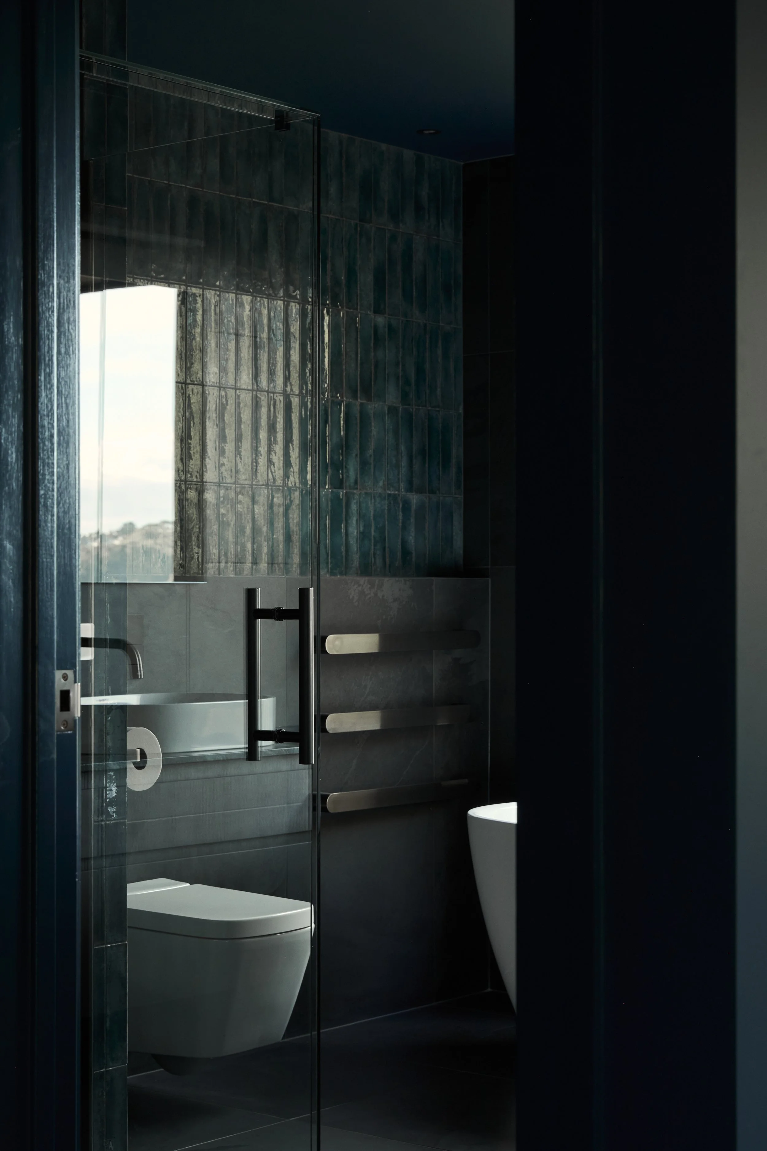 A modern bathroom with dark tiled walls, a glass shower door, a toilet, and a heated towel rack.